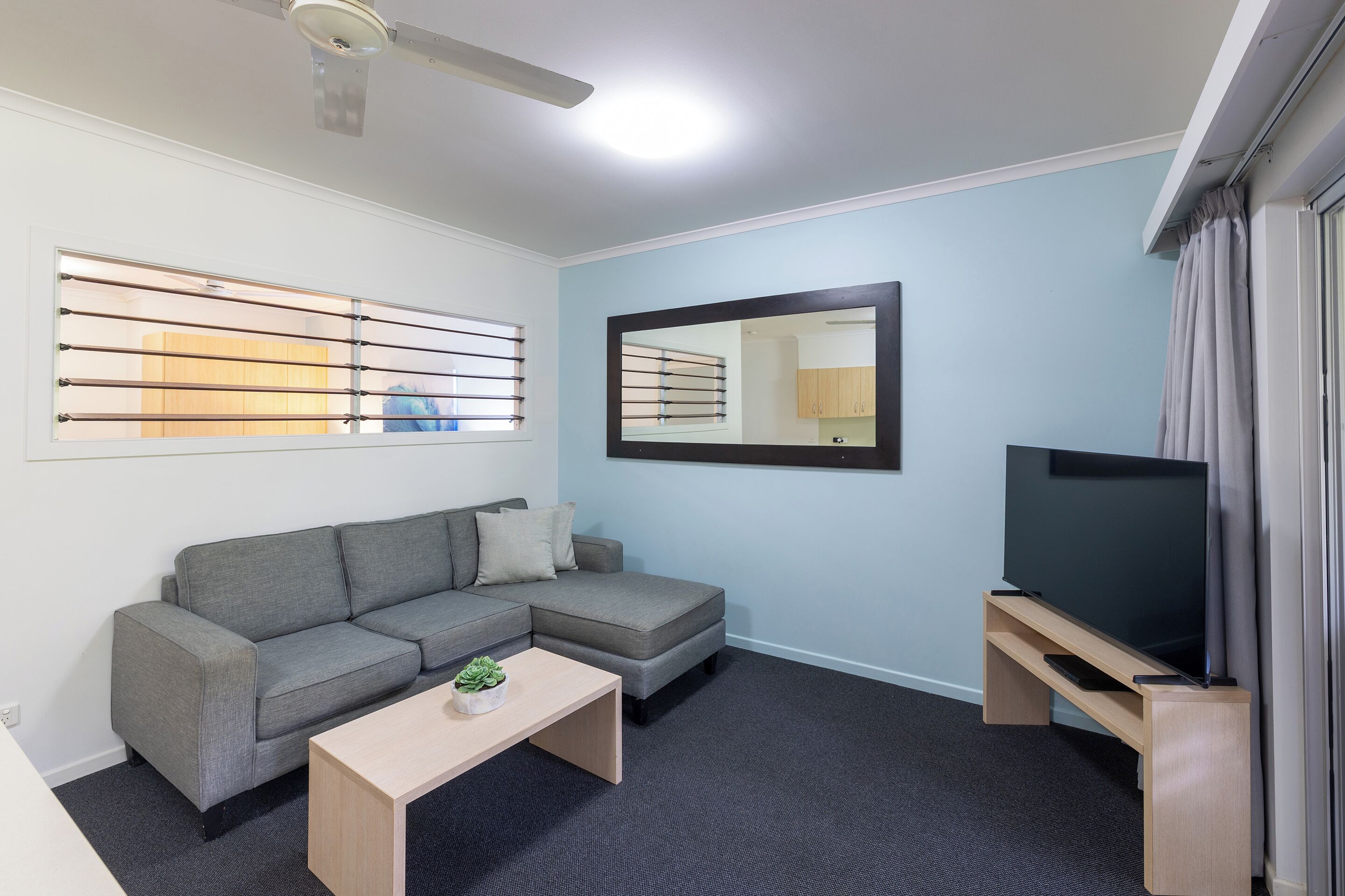 A modern living room with a sectional sofa, a coffee table, a flat-screen TV on a stand, a ceiling fan, and a window with blinds.