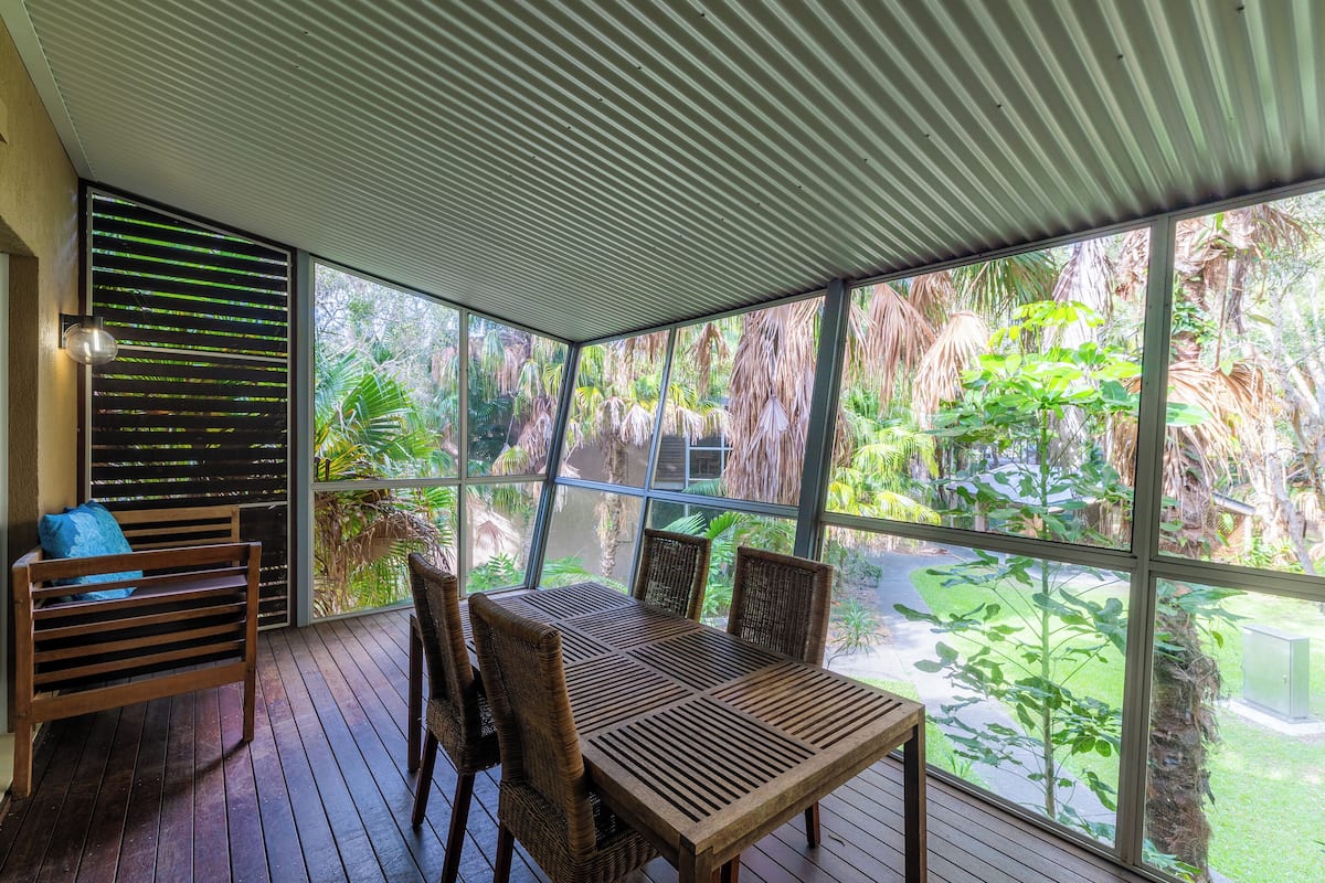 A covered patio with a wooden table and chairs, large windows, and a view of tropical vegetation.