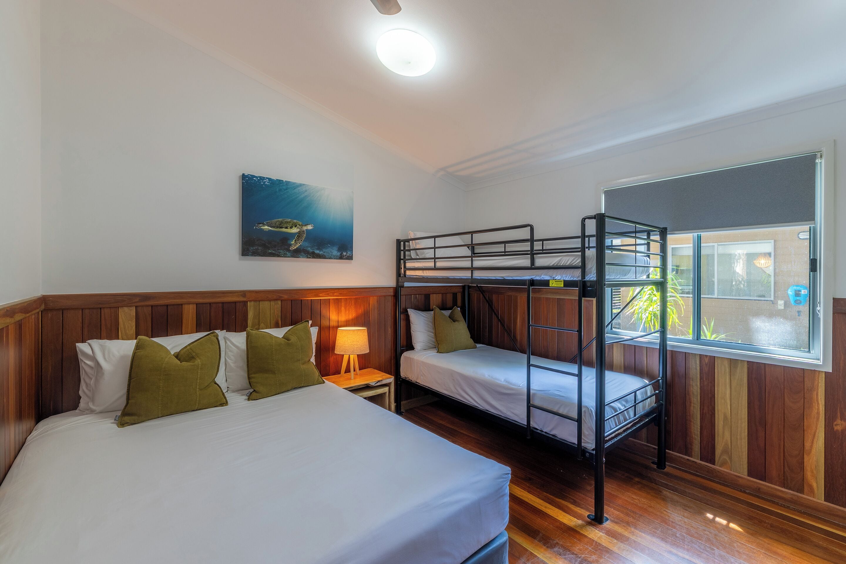 Una habitación con literas, un cabecero de madera, una mesita de noche con una lámpara y un cuadro en la pared.