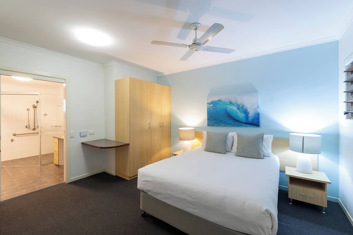 A hotel room with a bed, a ceiling fan, a nightstand, a lamp, a wooden wardrobe, a bathroom with a shower, and a wall painting.