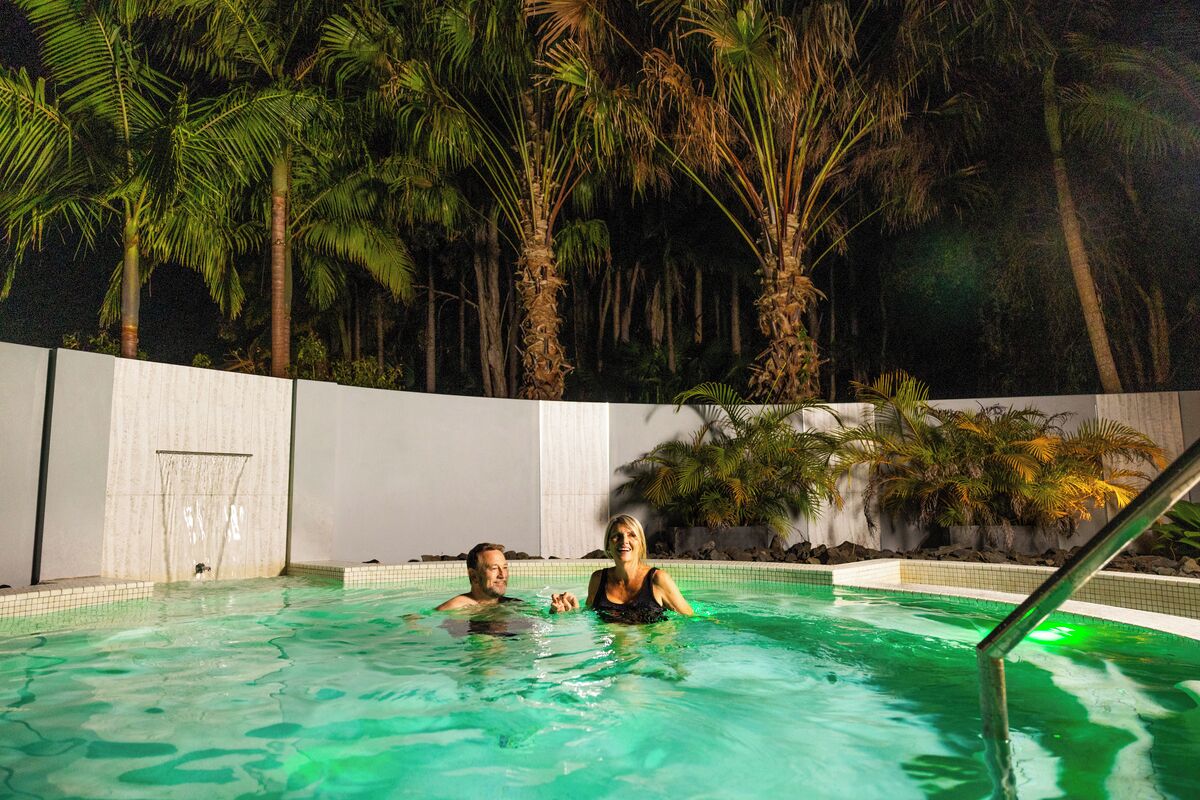 A swimming pool with two people, surrounded by palm trees and a white wall.