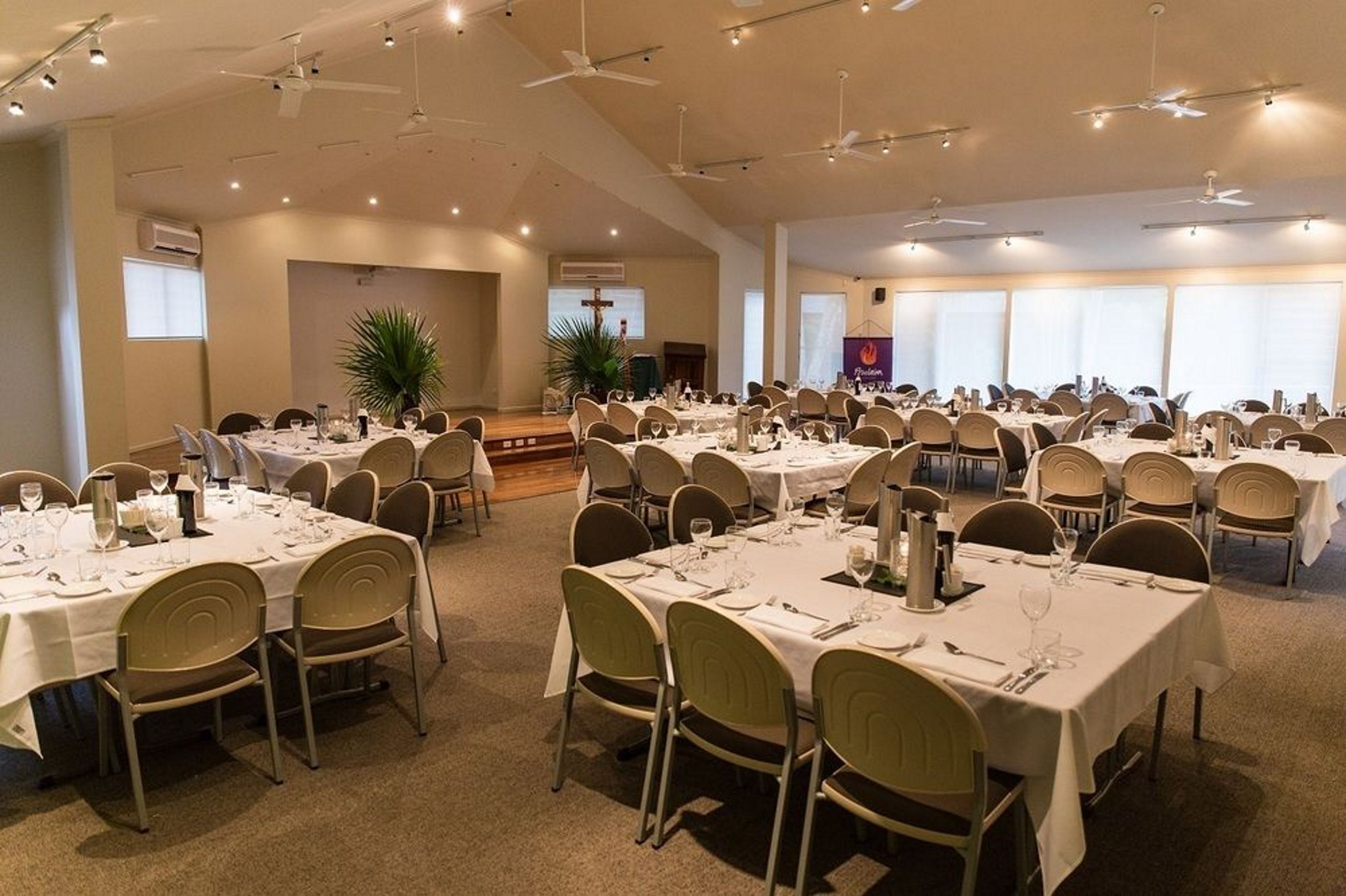 A banquet hall with multiple round tables set for dining, arranged in a spacious room with ceiling fans and recessed lighting.
