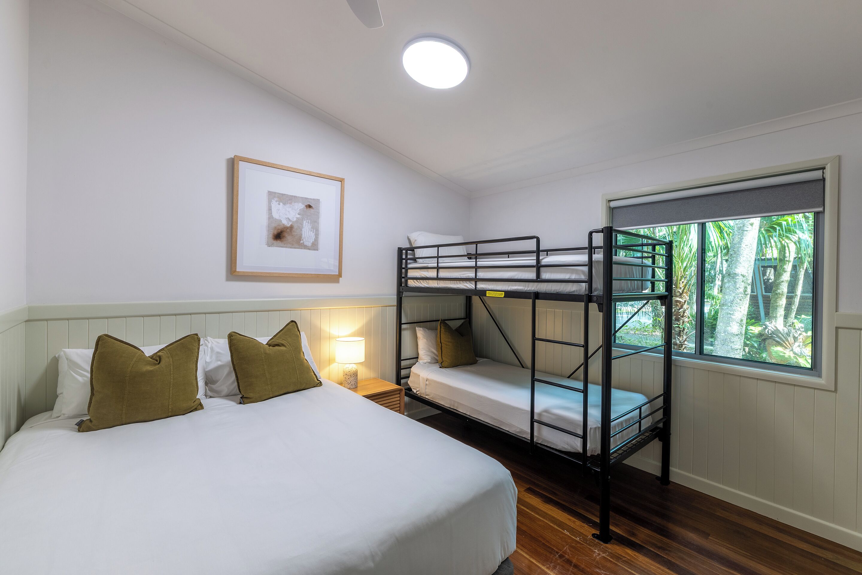 A room with bunk beds, a bedside table with a lamp, a framed picture on the wall, and a window with a view of greenery.