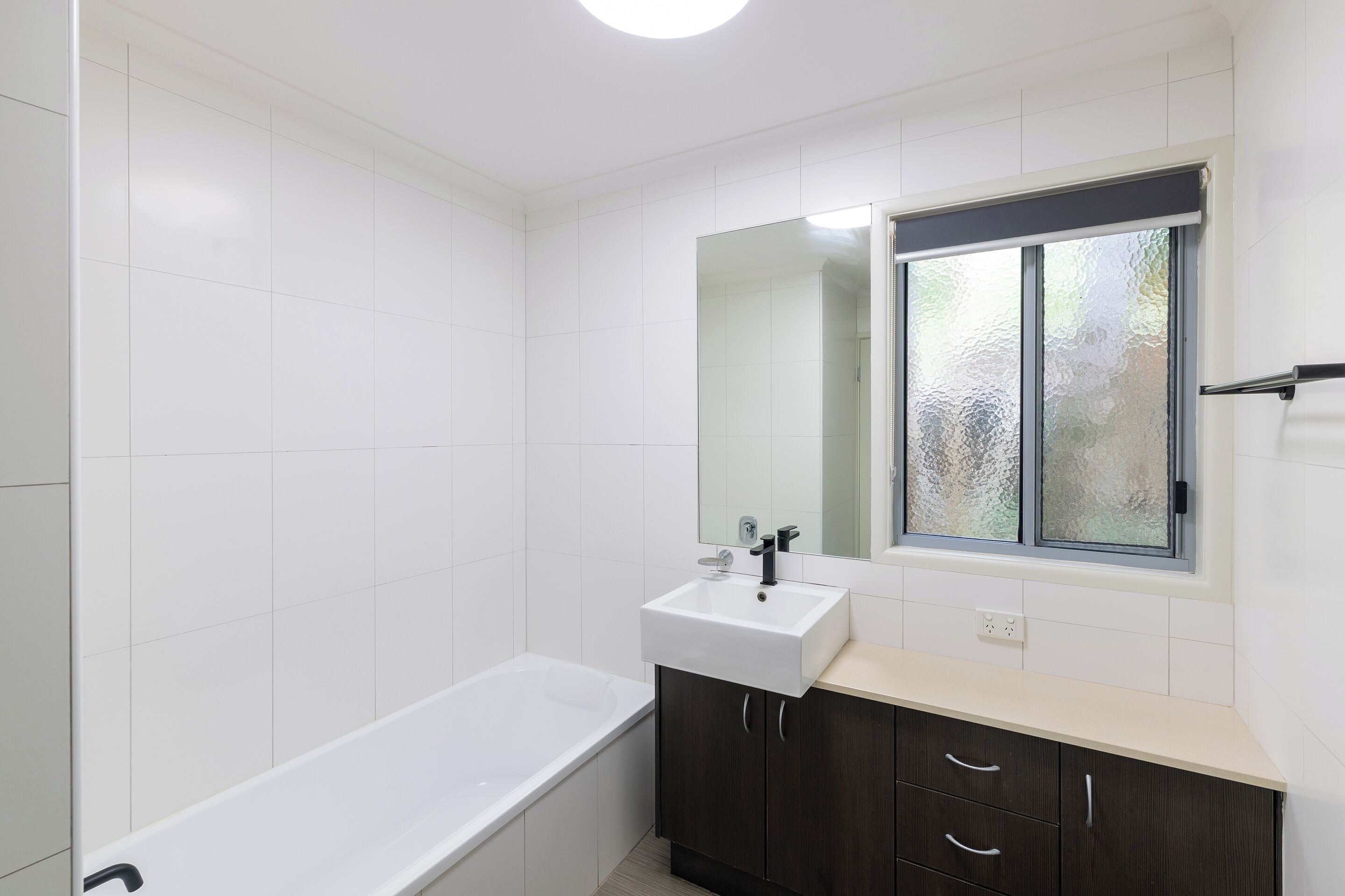 Et moderne badeværelse med et hvidt badekar, en vask med et spejl over og et vindue med matteret glas.