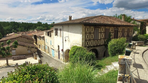 A traditional half-timbered building with a stone base and a tiled roof, surrounded by greenery and a cobblestone street. - VVF Gers Gascogne, Mauvezin (Mauvezin)