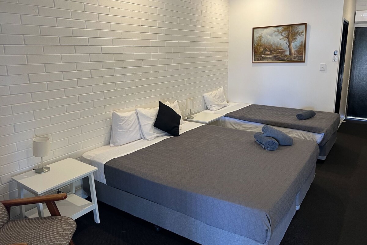 A hotel room with two beds, a white brick wall, a painting, and a small table with a lamp.