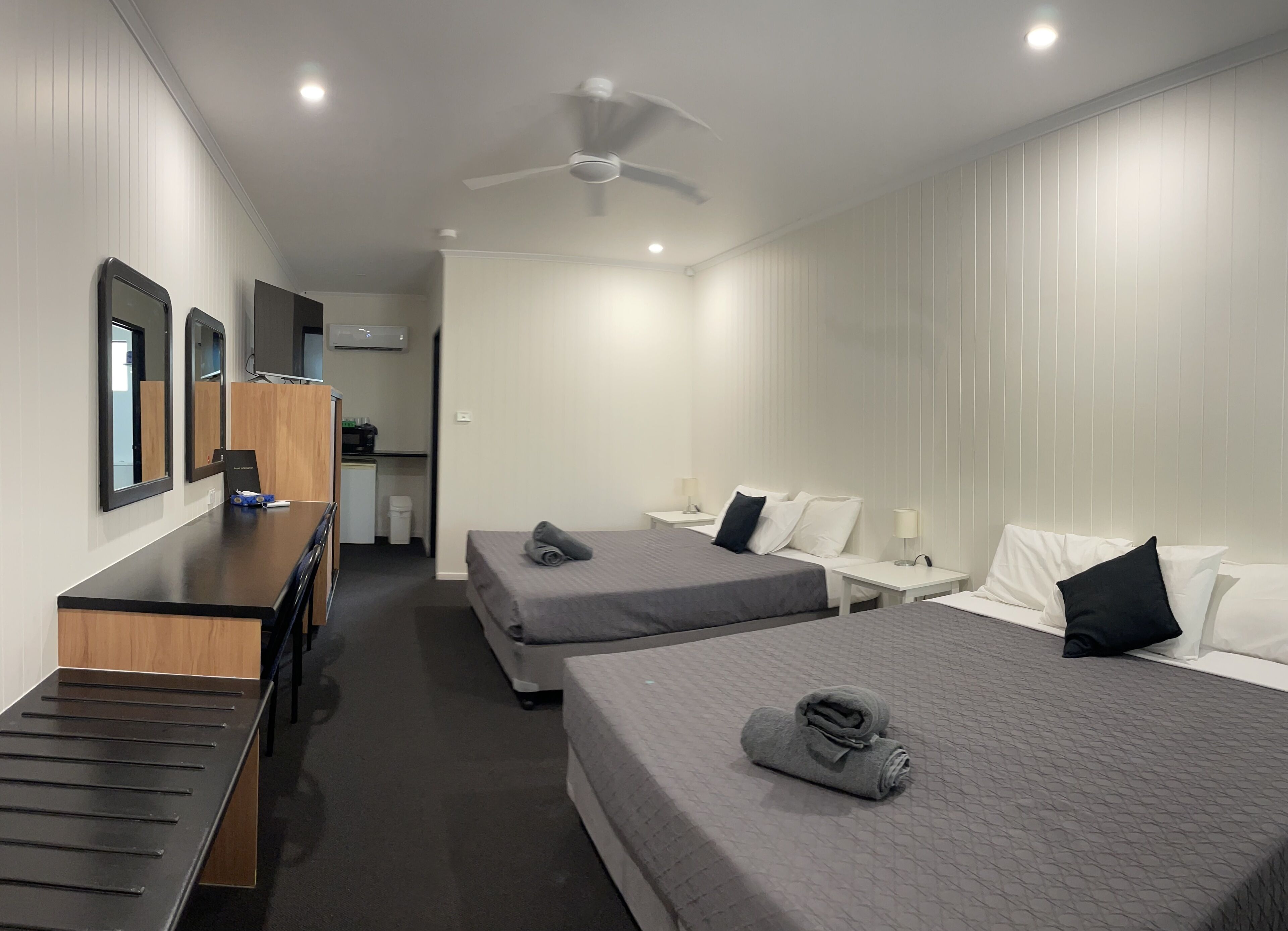 A hotel room with two beds, a desk, a chair, a wardrobe, and a ceiling fan.