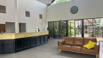 Uma área de recepção com um balcão identificado como "Albert Park Motel", um relógio e um sofá de couro marrom com uma almofada amarela.