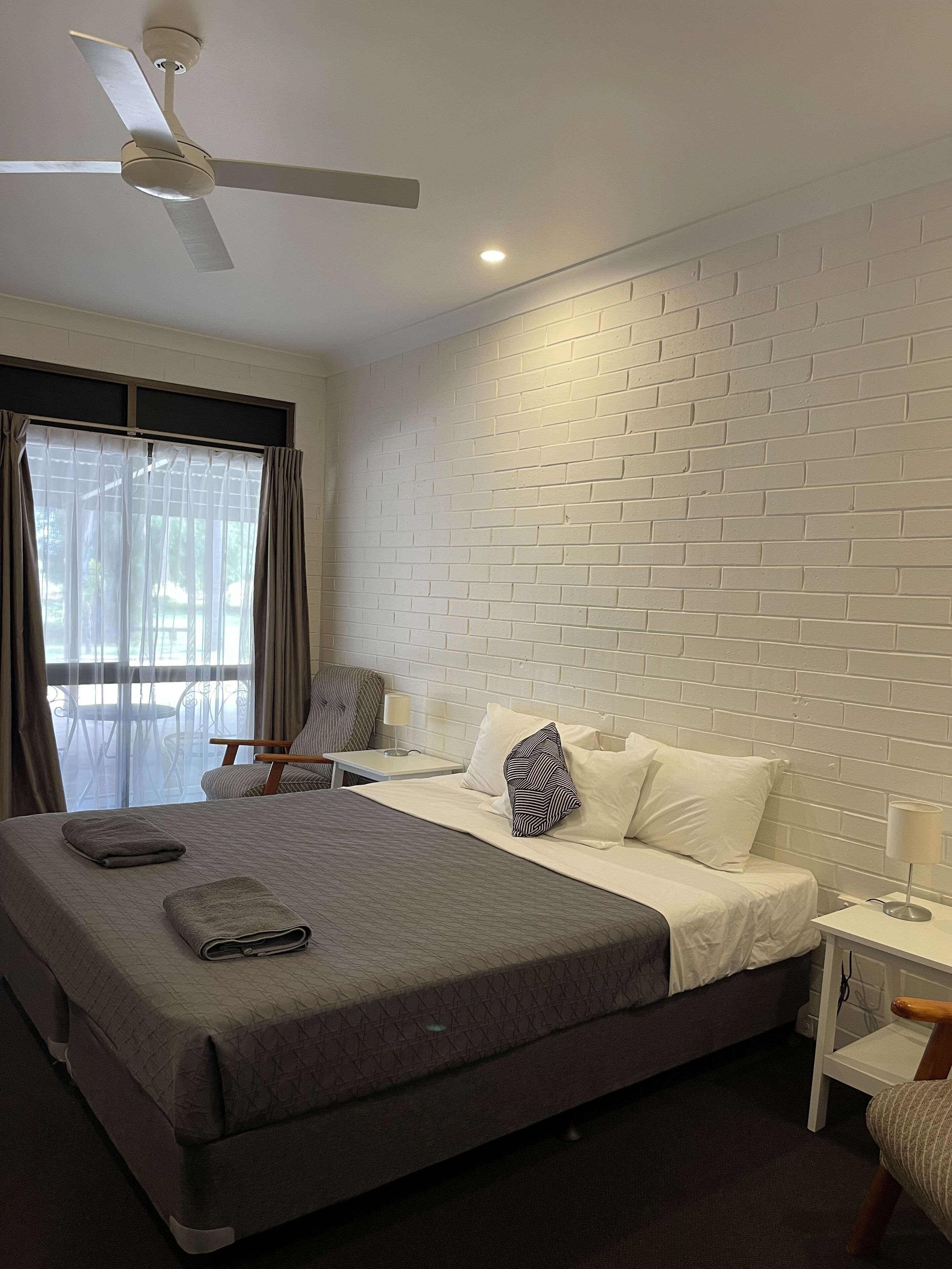 A hotel room with a large bed, a ceiling fan, a brick-patterned wall, a window with curtains, and a small table with a lamp.