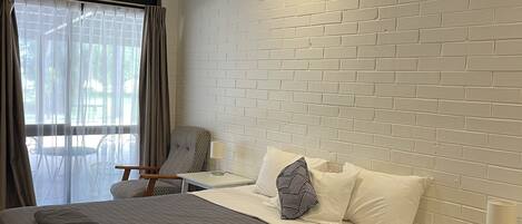 A hotel room with a large bed, a ceiling fan, a brick-patterned wall, a window with curtains, and a small table with a lamp.