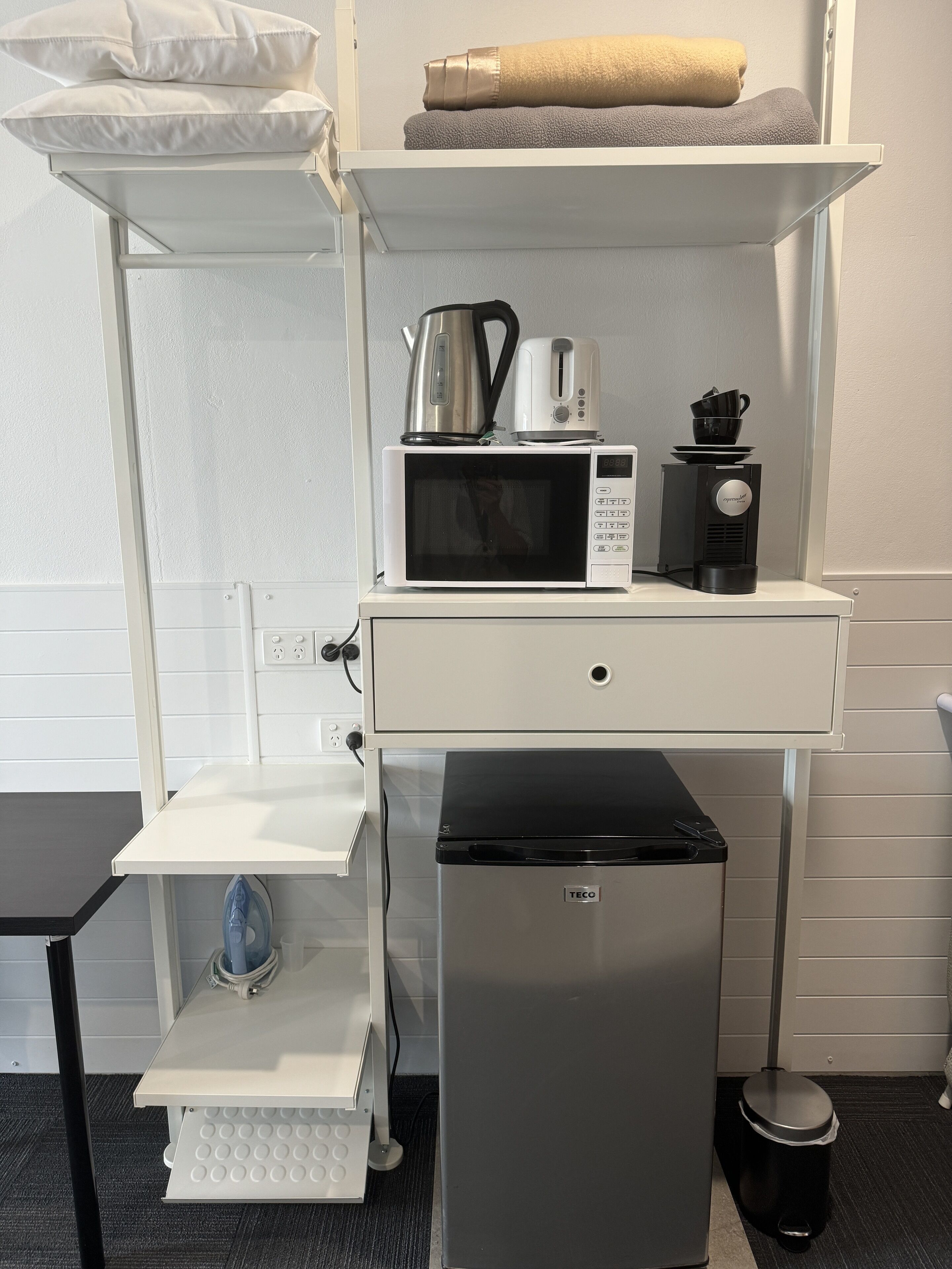 A compact kitchenette with a microwave, kettle, and coffee maker on a white countertop, a small refrigerator, and a trash bin.