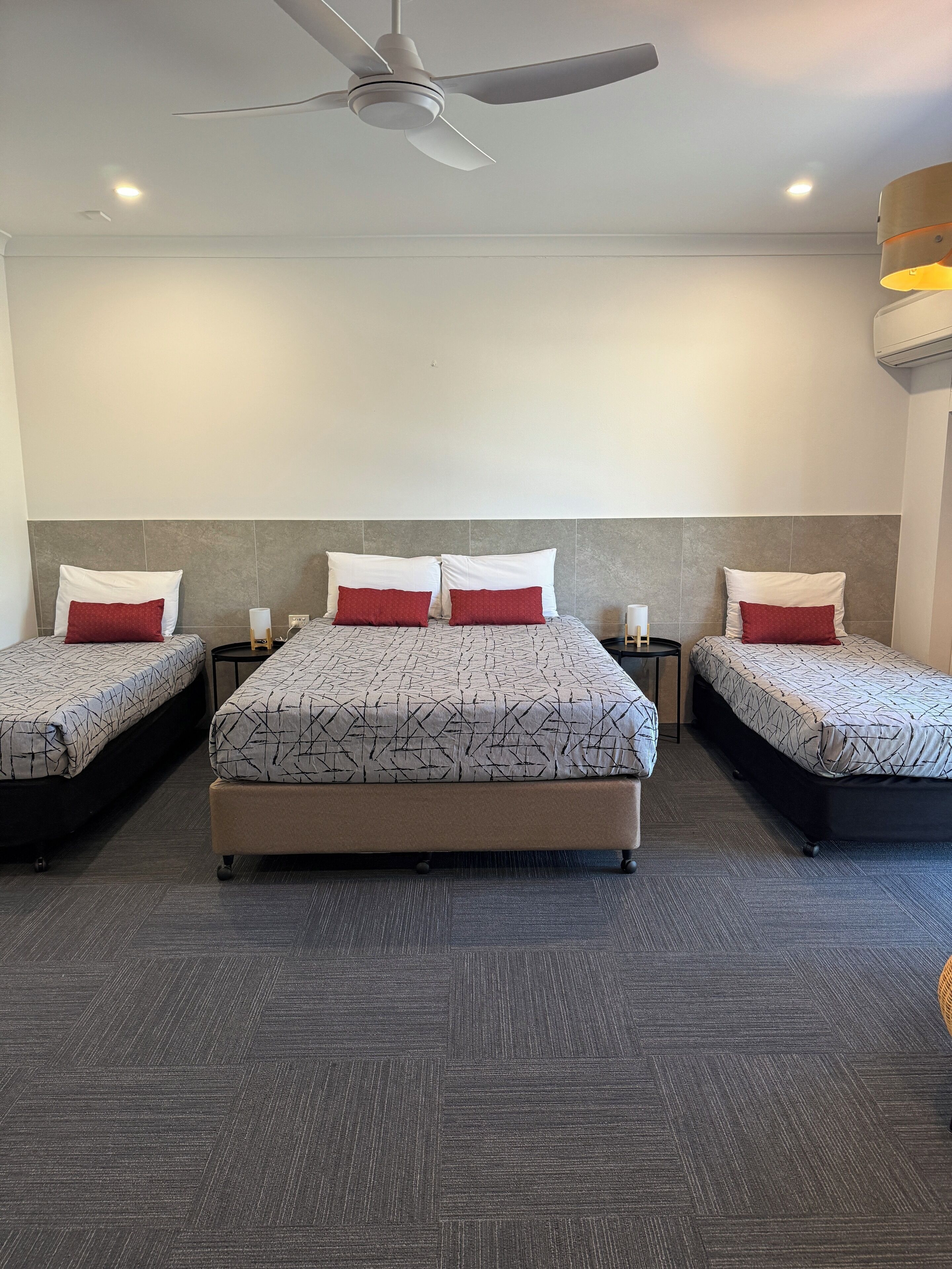 A hotel room with a large bed, two smaller beds, a ceiling fan, and a nightstand with a lamp.