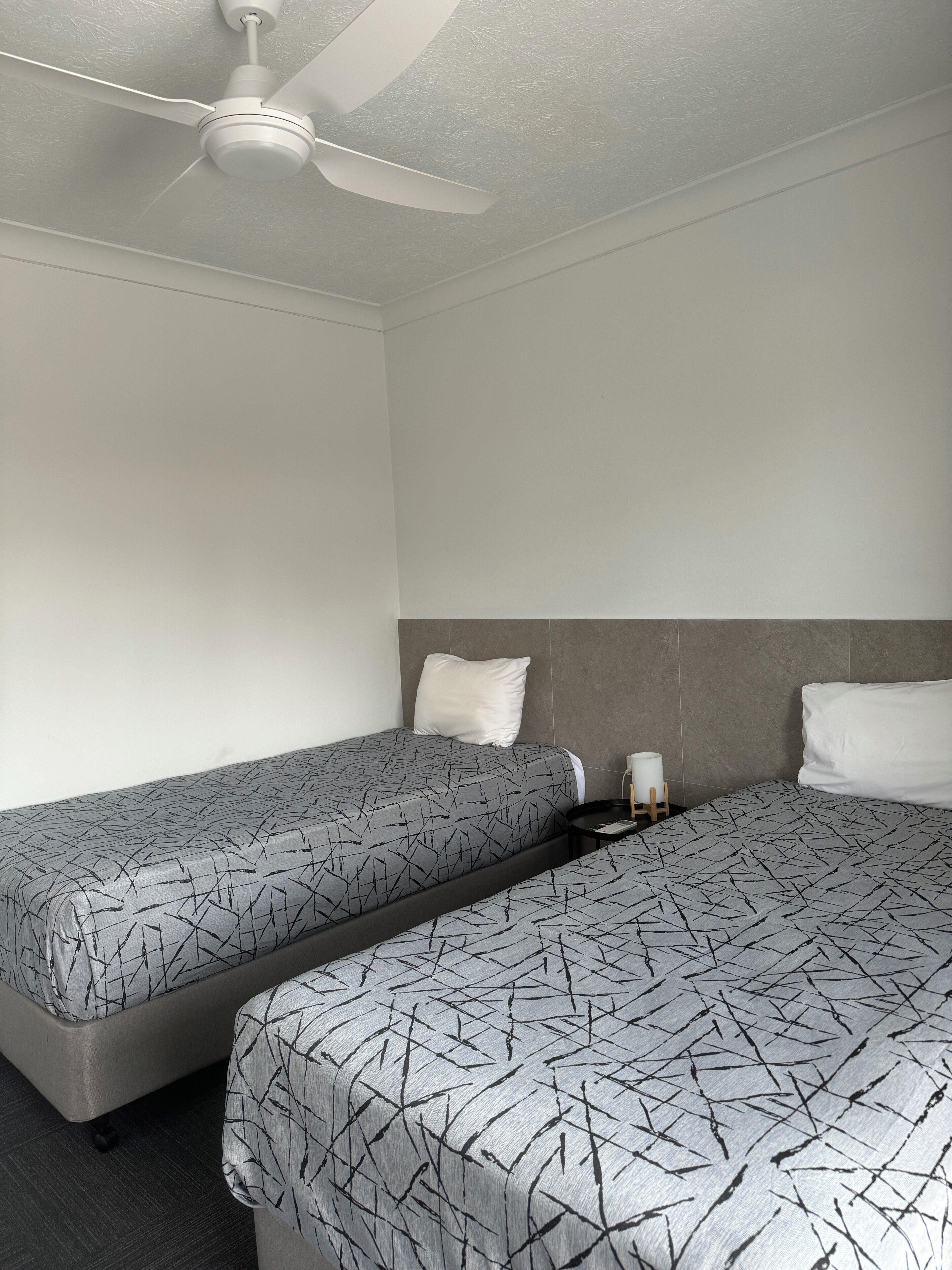 Una habitación de hotel con dos camas individuales, un ventilador de techo y una mesita de noche pequeña con una lámpara.