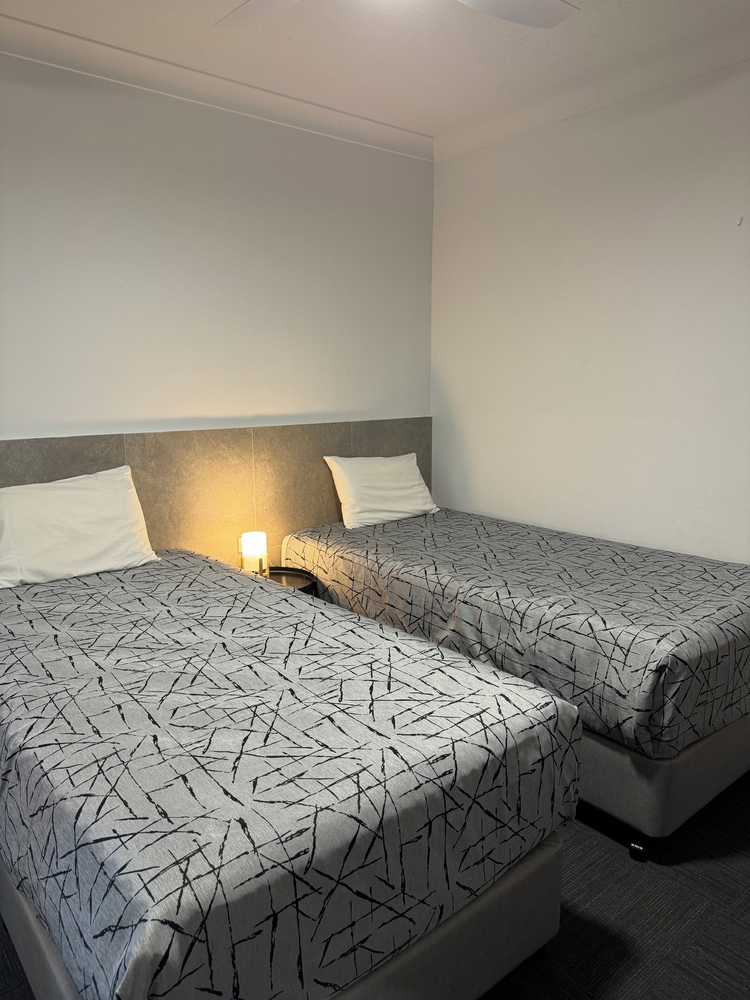 A hotel room with two beds, each with a patterned bedspread, a bedside table with a lamp, and a plain wall.