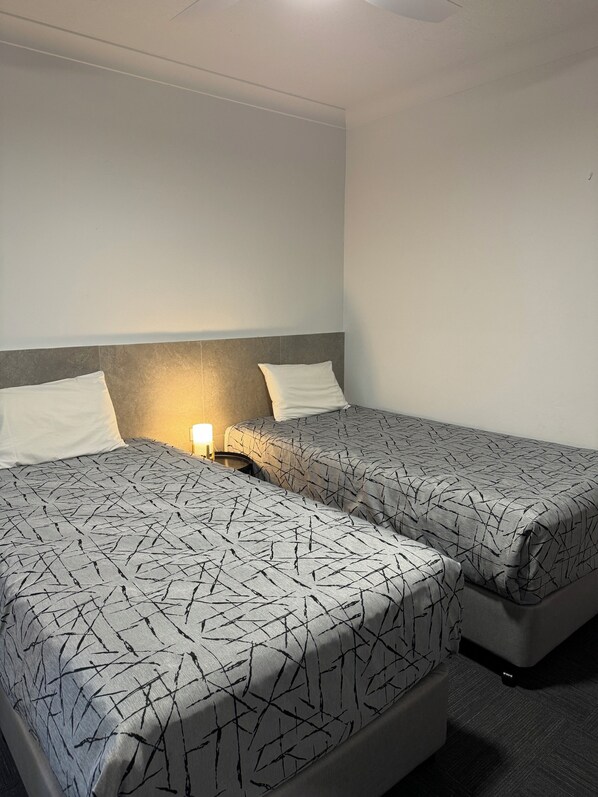 A hotel room with two beds, each with a patterned bedspread, a bedside table with a lamp, and a plain wall.