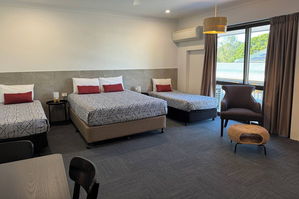 Habitación de hotel con una cama grande, dos camas más pequeñas, una silla, una mesa y un ventilador de techo.