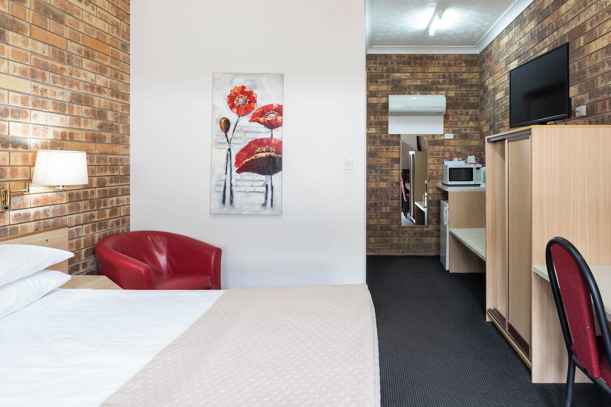 Habitación de hotel con pared de ladrillo, un cuadro de flores, una cama, una silla roja, un escritorio, un televisor y un microondas.