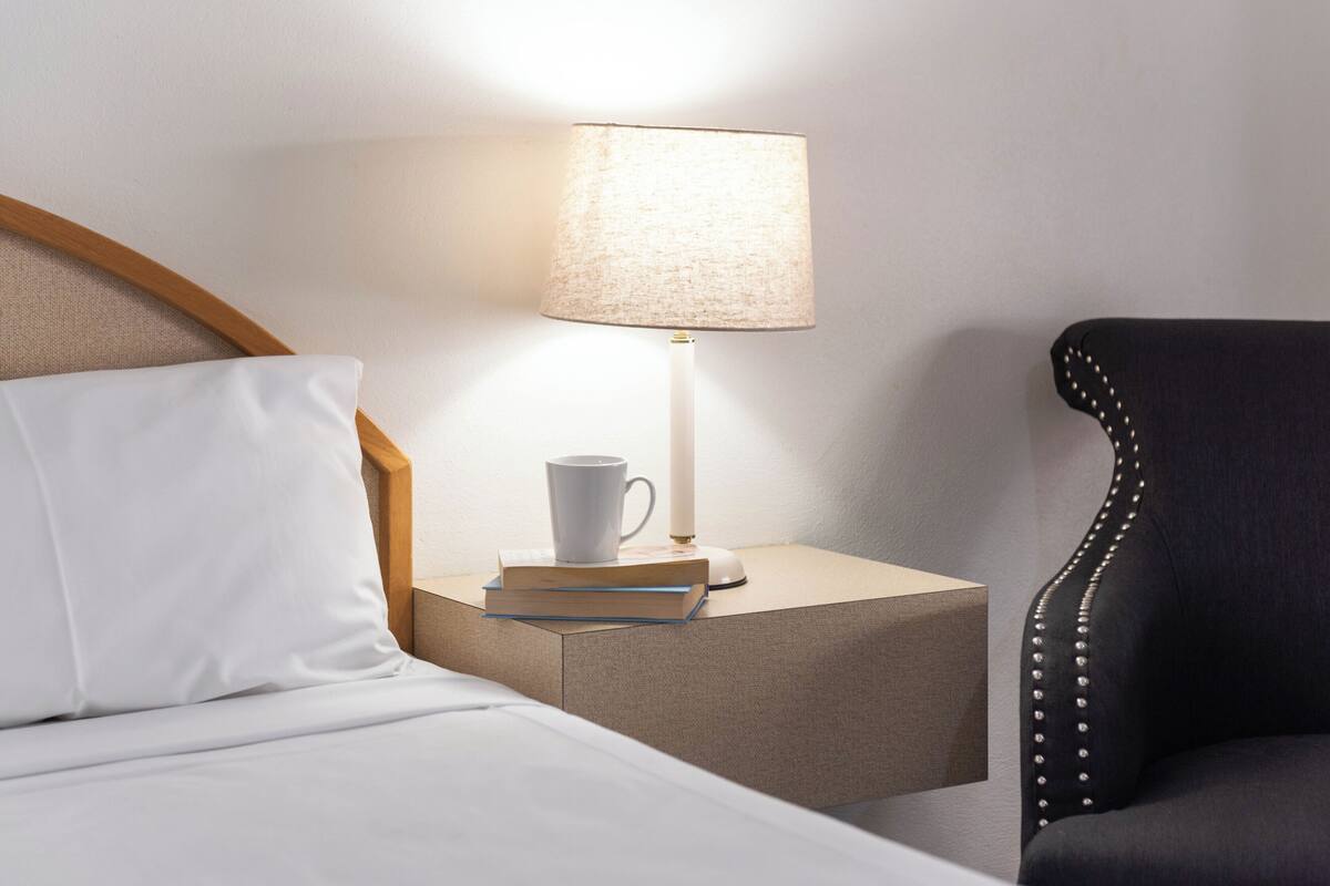 Una cama bien hecha con un almohadón blanco, una mesita de noche con una lámpara, una taza blanca y un libro.