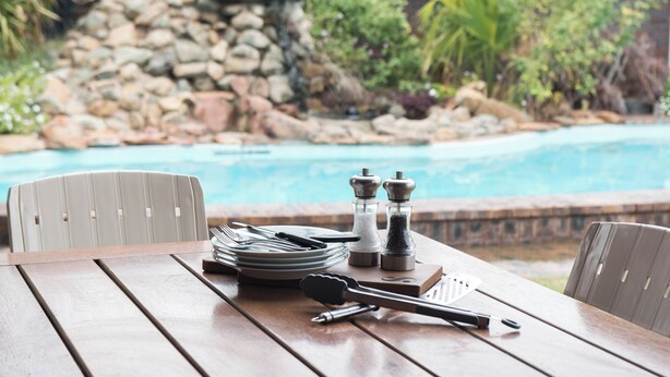 Une table en bois, dressée avec des assiettes, des couverts et des sali ères et poivrières, donnant sur une piscine, avec un mur de pierre et de la verdure en arrière-plan.