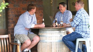 Trois hommes sont assis à une table en bois, près d’une barrique, et tiennent chacun une bouteille de bière.