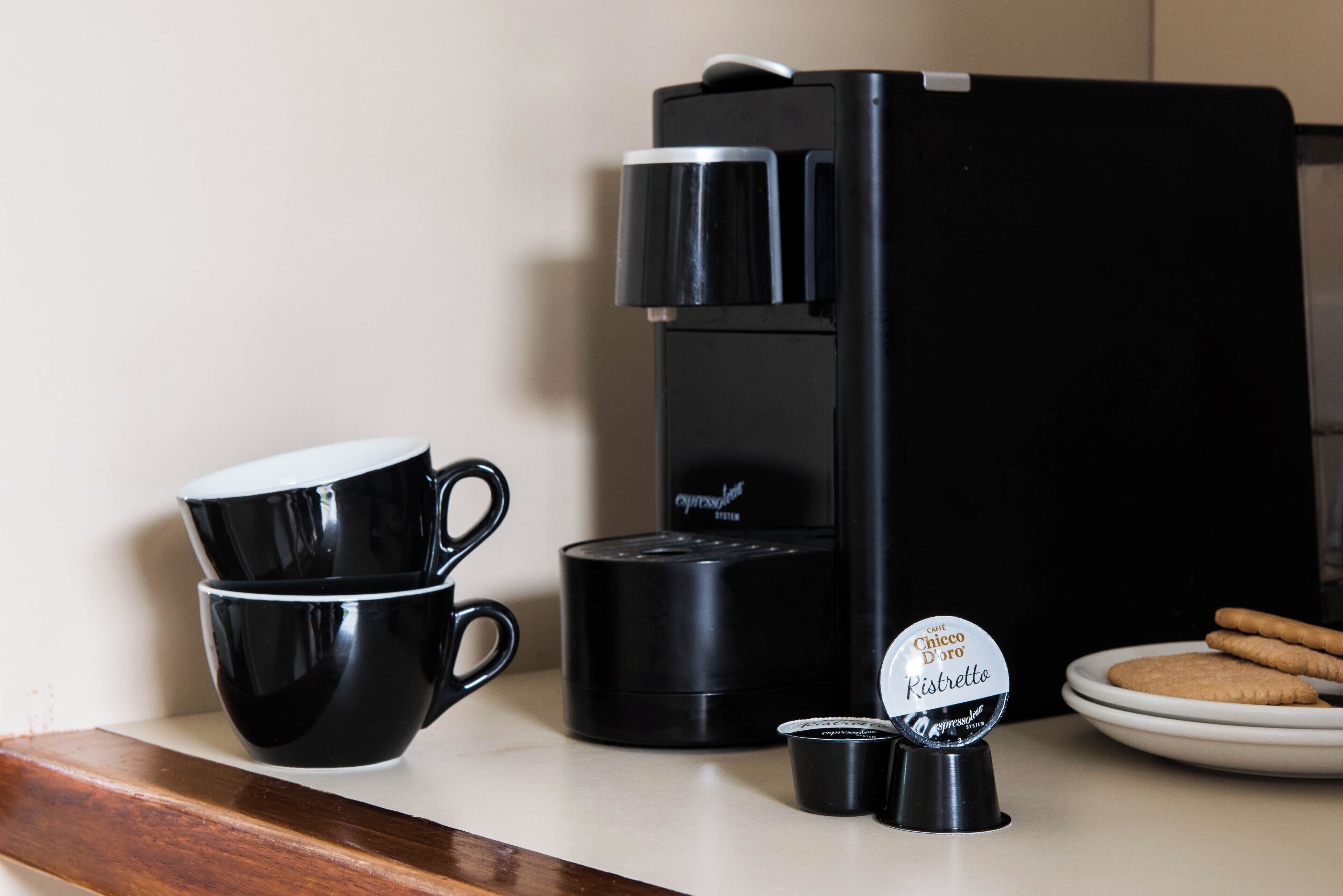 A coffee maker, two black cups, a plate with cookies, and a coffee pod on a countertop.