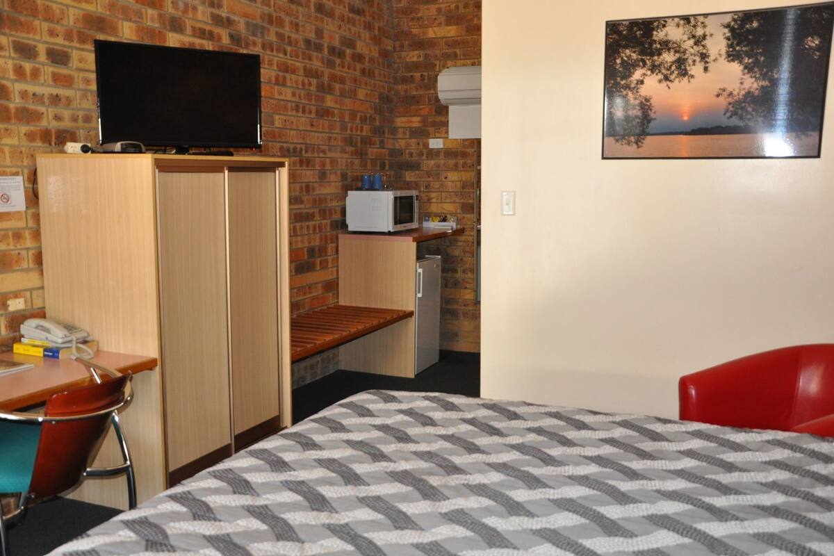 Una habitación con cama, escritorio, silla, televisor, microondas y un cuadro en la pared.