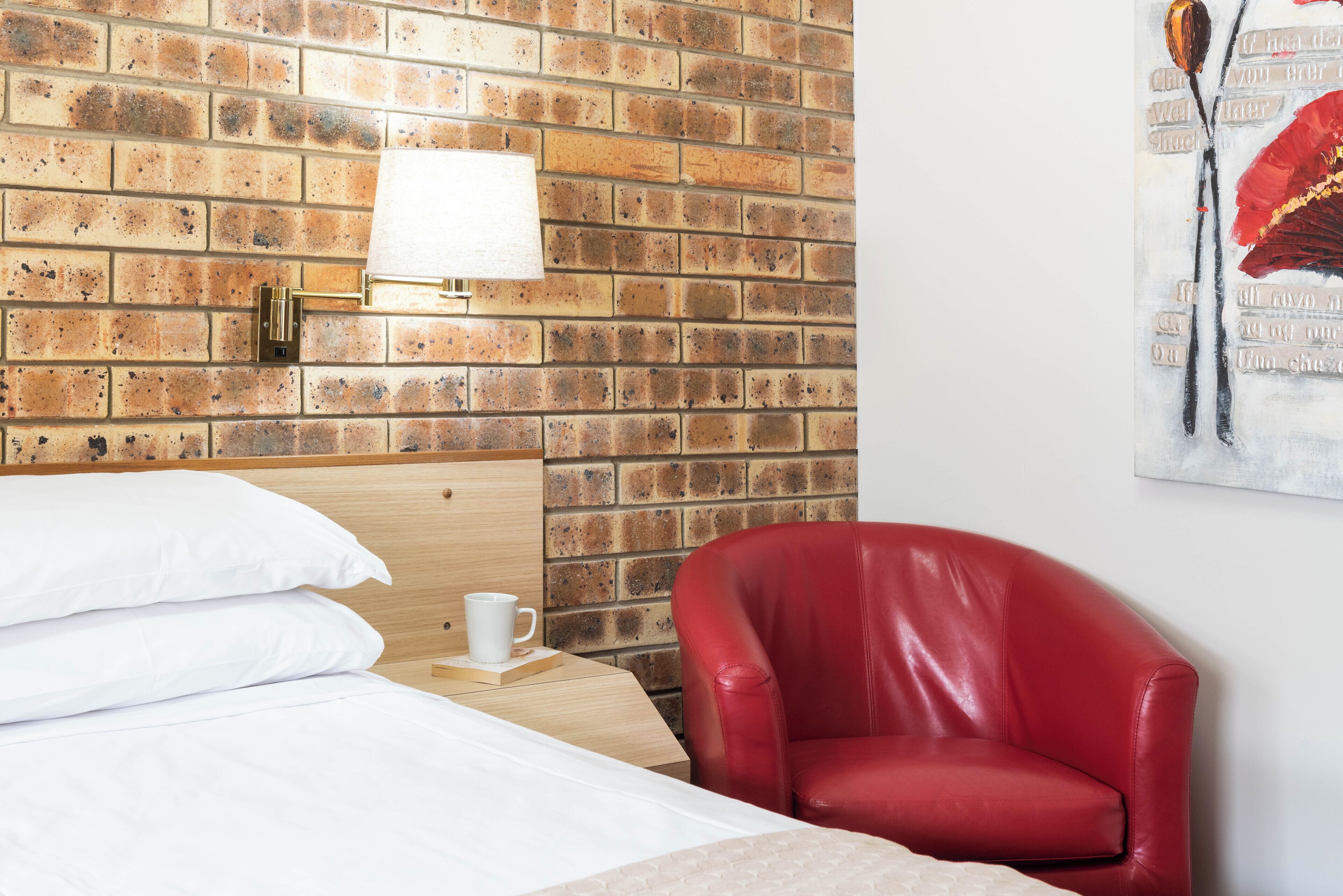 Ein Hotelzimmer mit einer Wand im Ziegelsteinmuster, einem hölzernen Kopfteil, einem roten Ledersessel, einem weißen Bett mit Kissen und einer weißen Tasse auf einem Nachttisch.