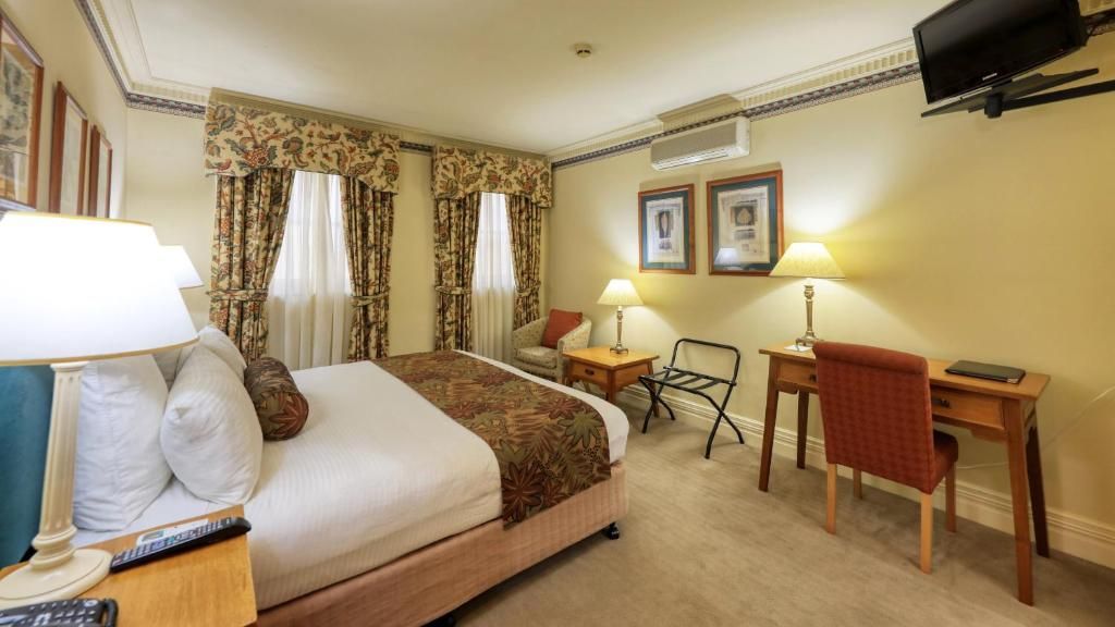 Quarto de hotel com uma cama, duas mesas de cabeceira, escrivaninha, cadeira, televisão e cortinas.