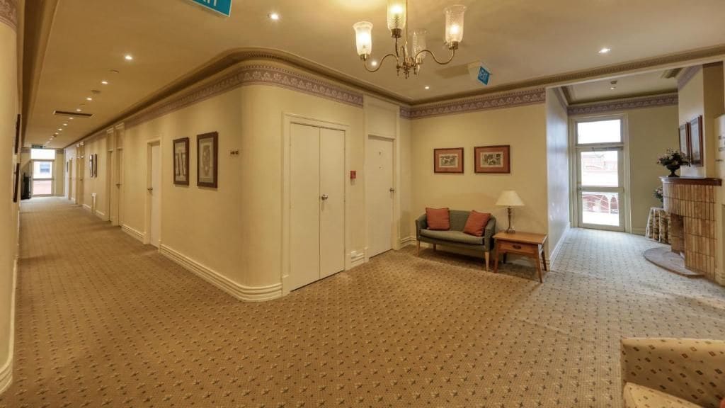 A hotel corridor with a patterned carpet, a sofa with cushions, a lamp, and framed pictures on the walls.