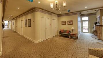 A hotel corridor with a patterned carpet, a sofa with cushions, a lamp, and framed pictures on the walls.