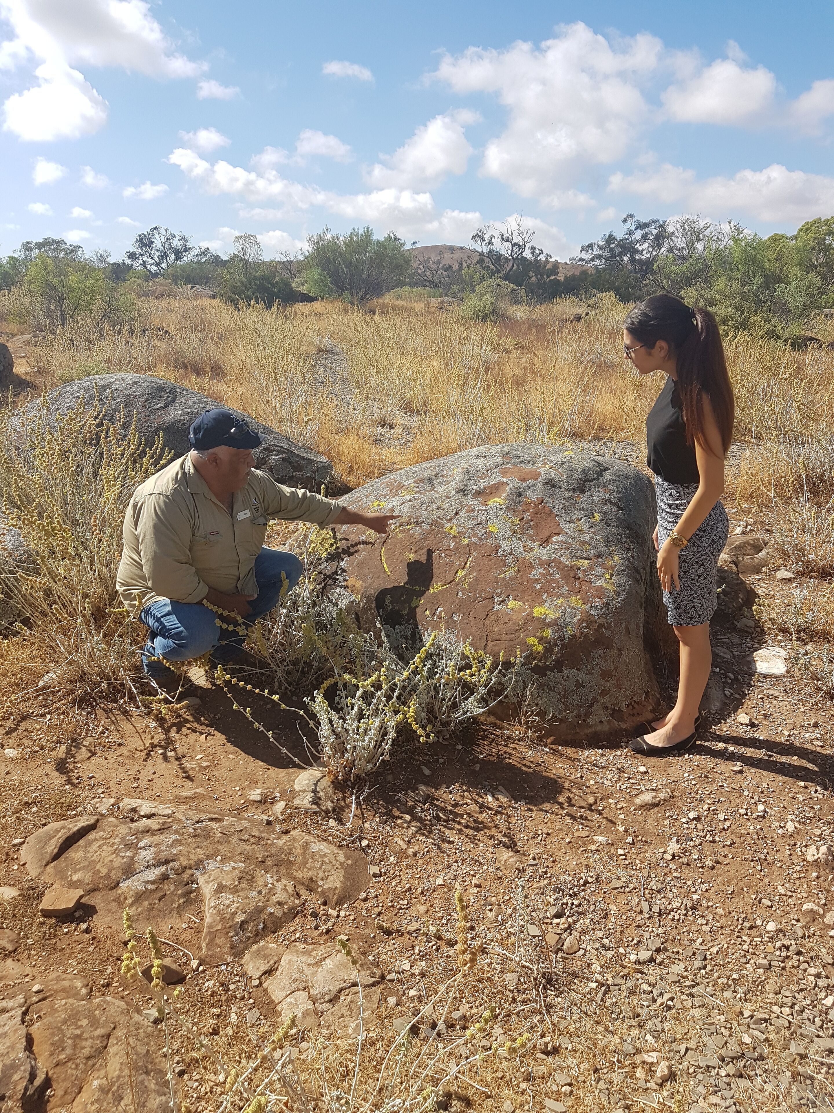 Dos personas observan una roca grande en un paisaje seco y rocoso, con poca vegetación y colinas al fondo.