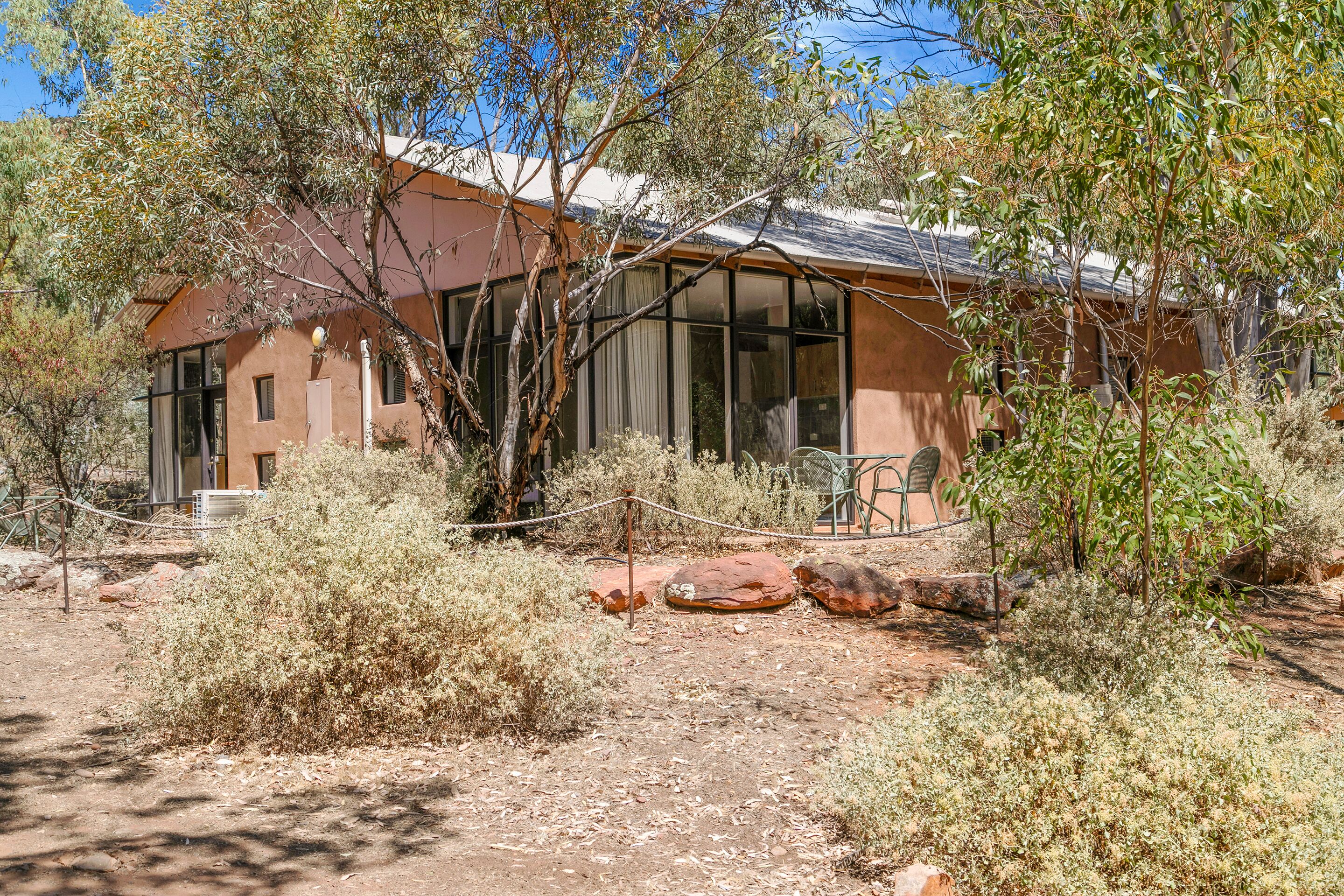A rustic building with large windows, surrounded by dry shrubs and rocks.