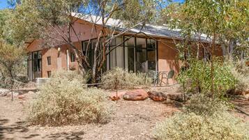 A rustic building with large windows, surrounded by dry shrubs and rocks.