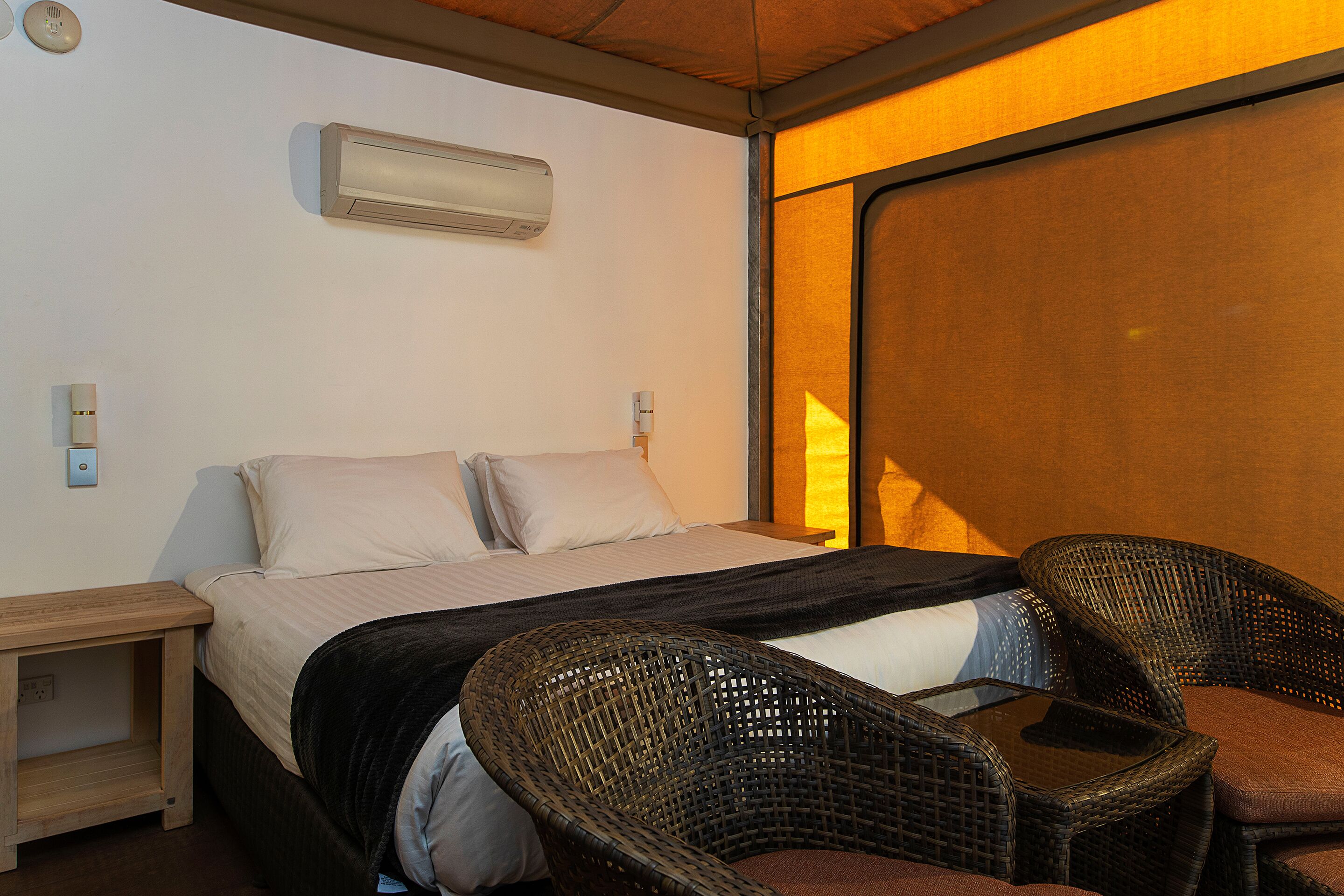 A hotel room with a bed, two wicker chairs, a nightstand, and an air conditioning unit on the wall.