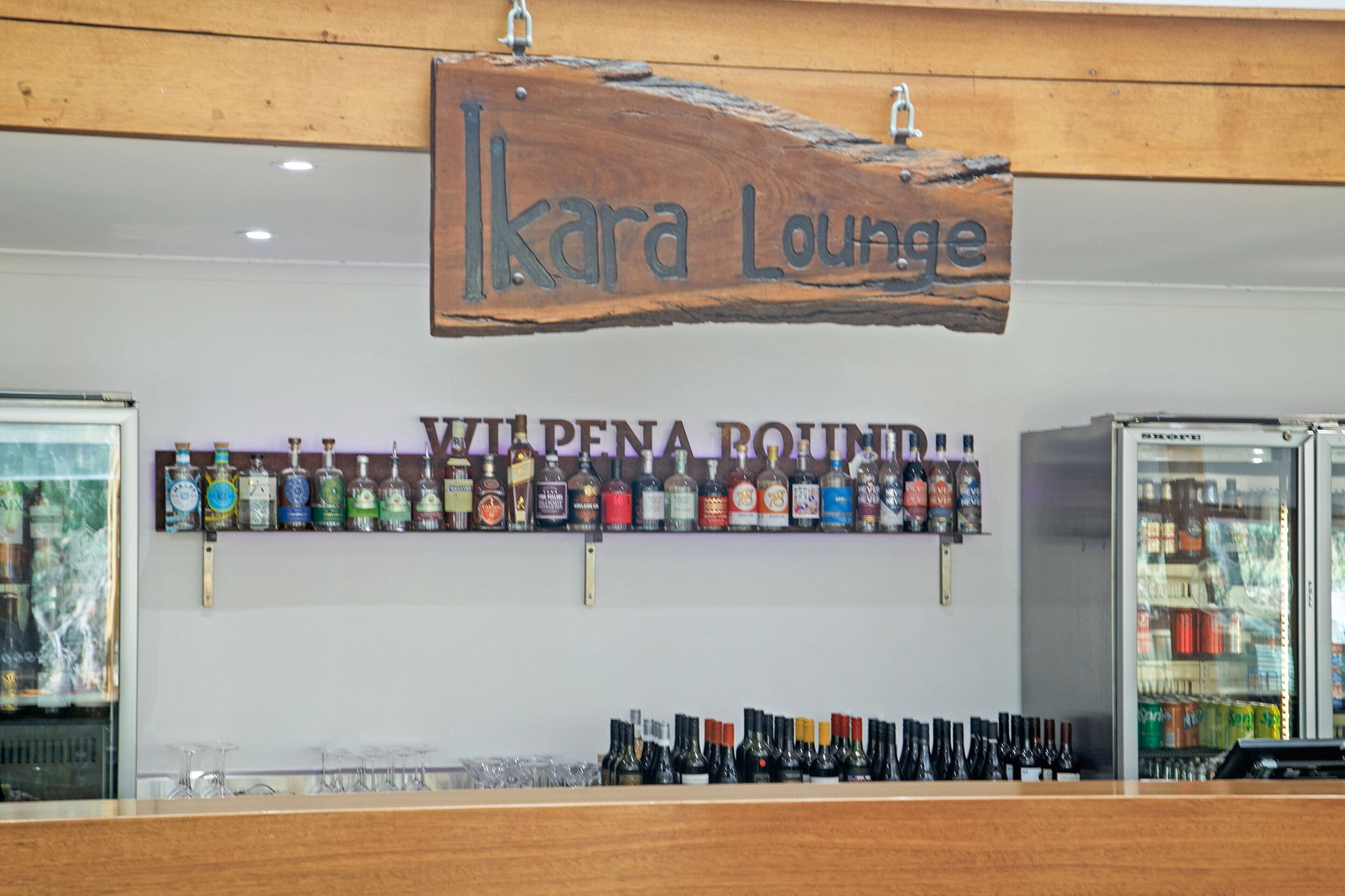 En bardisk med en träskylt där det står ”Ikara Lounge” och en hylla med olika sorters spritflaskor.