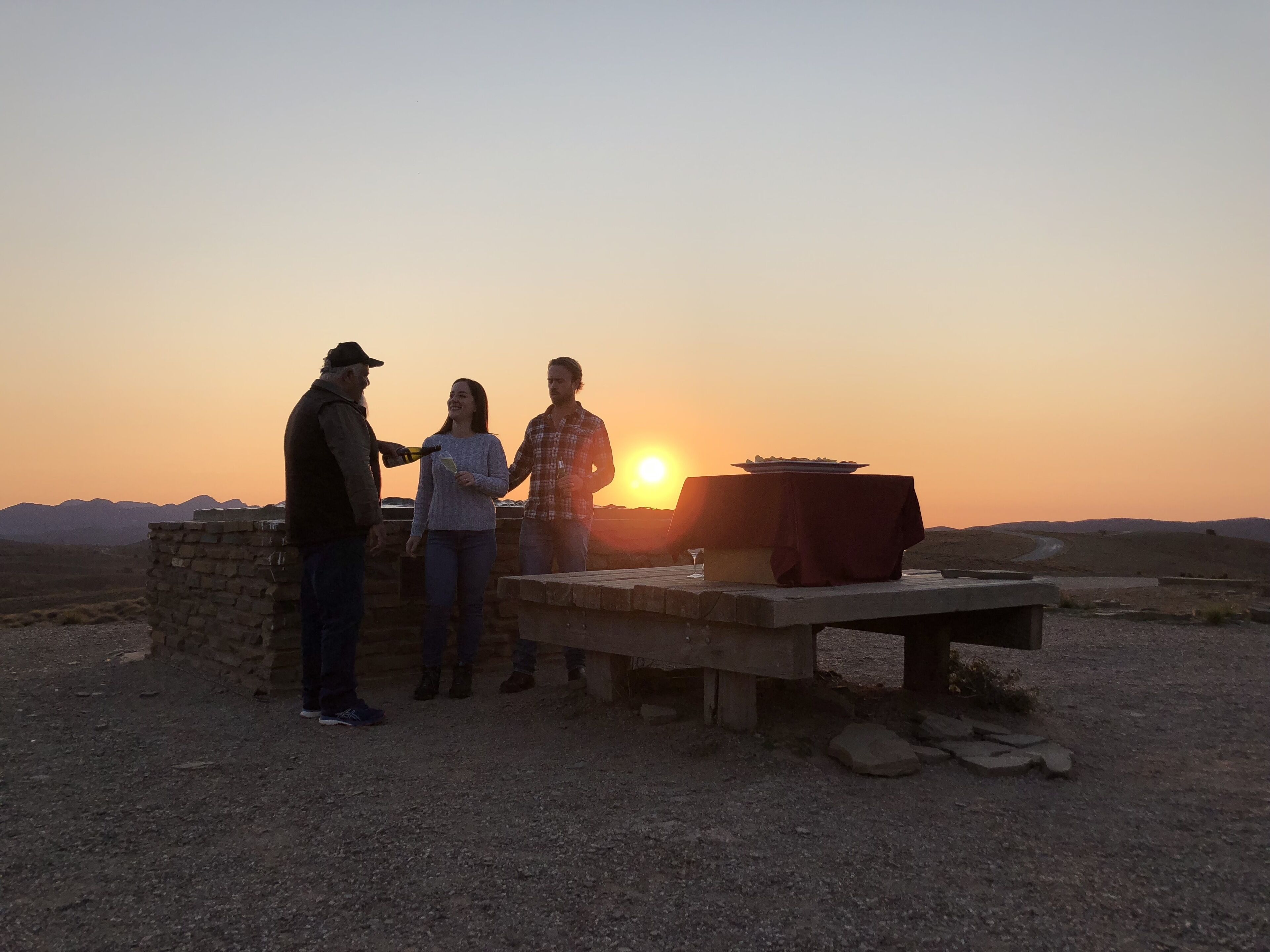 Three people standing near a table with a plate and a glass, with a sunset in the background.