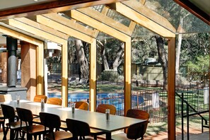 An outdoor dining area with wooden tables and chairs, a glass enclosure, and a view of a swimming pool and trees.