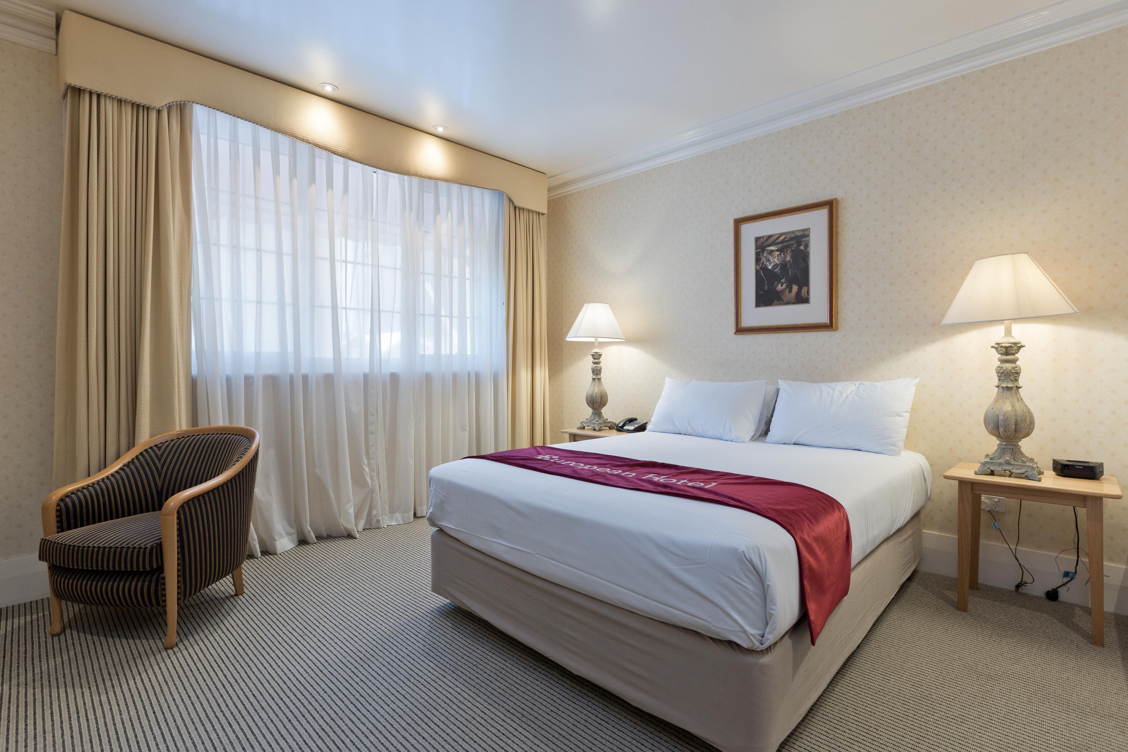 Una habitación de hotel con una cama grande, una silla, dos mesitas de noche con lámparas y una ventana con cortinas.