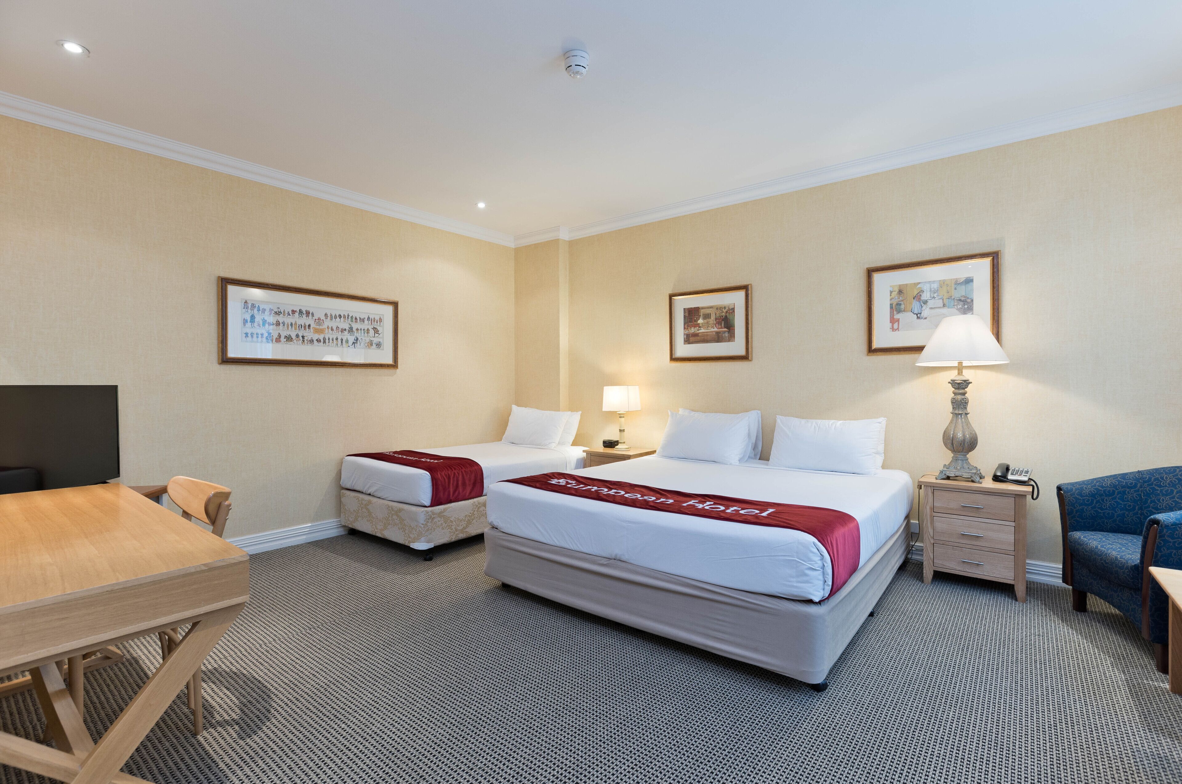 Una habitación de hotel con dos camas, un escritorio, una silla, una lámpara y cuadros en la pared.