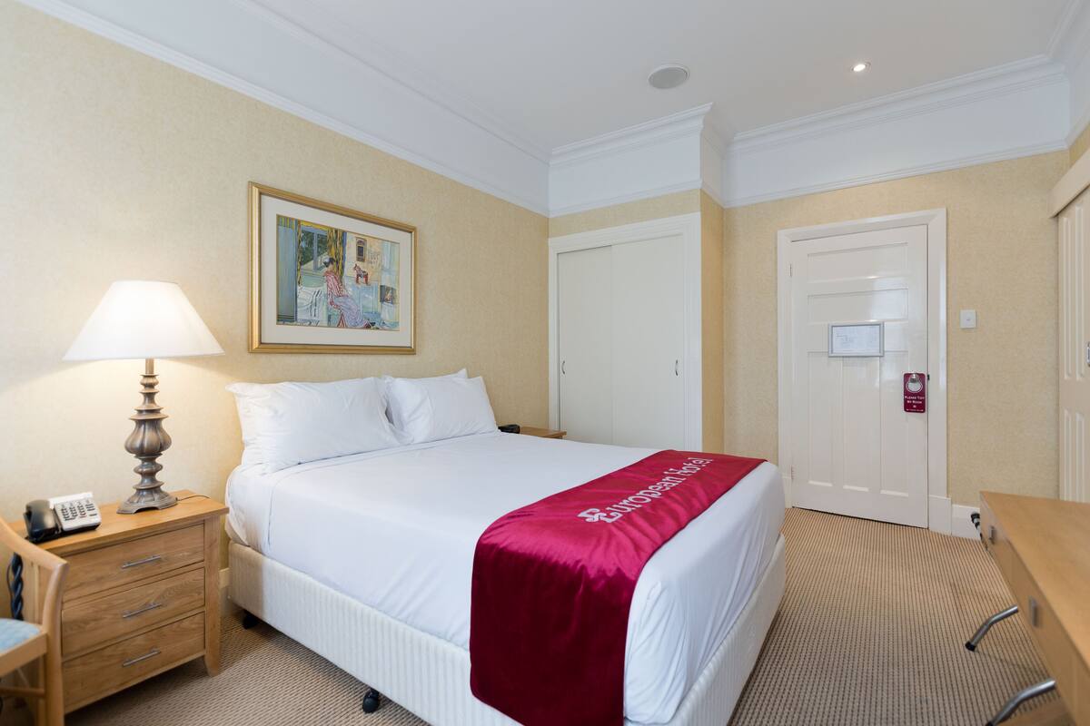 A hotel room with a neatly made bed, a bedside lamp, a desk, and a door with a sign.
