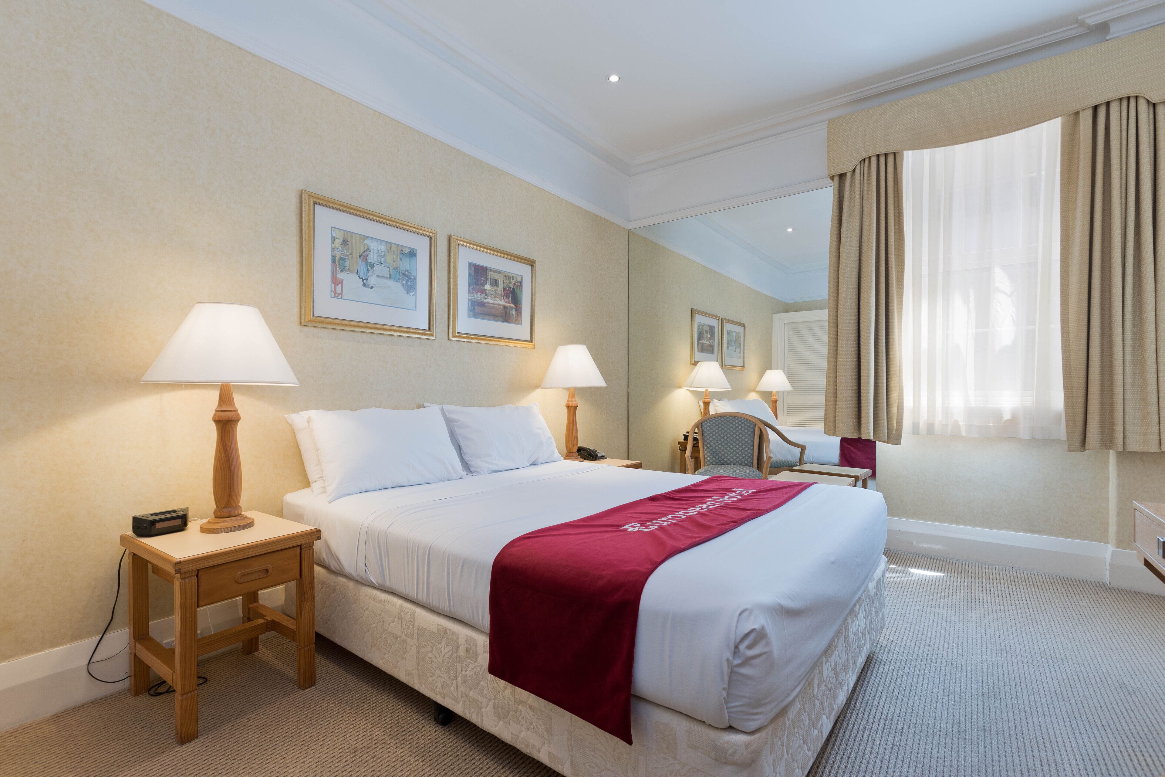 Habitación de hotel con una cama grande, dos lámparas de noche, una mesita de noche, una silla y un amplio ventanal con cortinas.