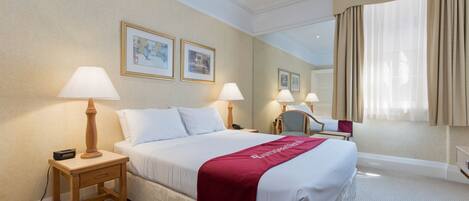 Quarto de hotel com uma cama grande, duas luminárias de cabeceira, mesa de cabeceira, uma cadeira e uma janela ampla com cortinas.