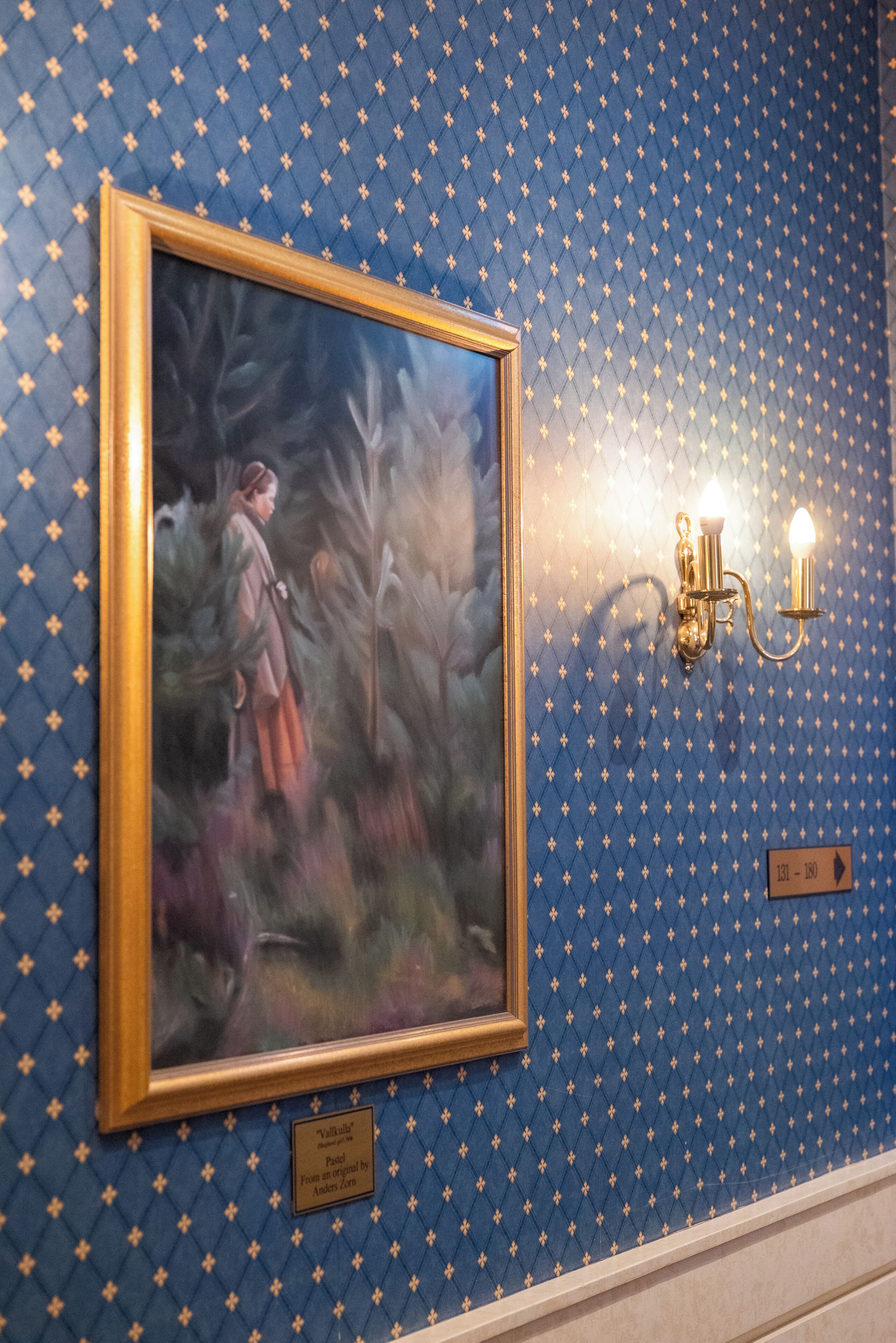 Een lijst met een schilderij aan de muur, met een blauwe achtergrond met patroon, en een wandlamp.