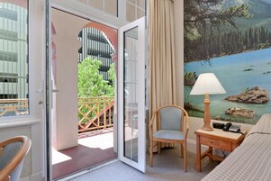 Habitación de hotel con puerta corrediza de vidrio que da a un balcón, una cama, una silla, una mesita de noche con lámpara, y un cuadro de un lago y montañas.