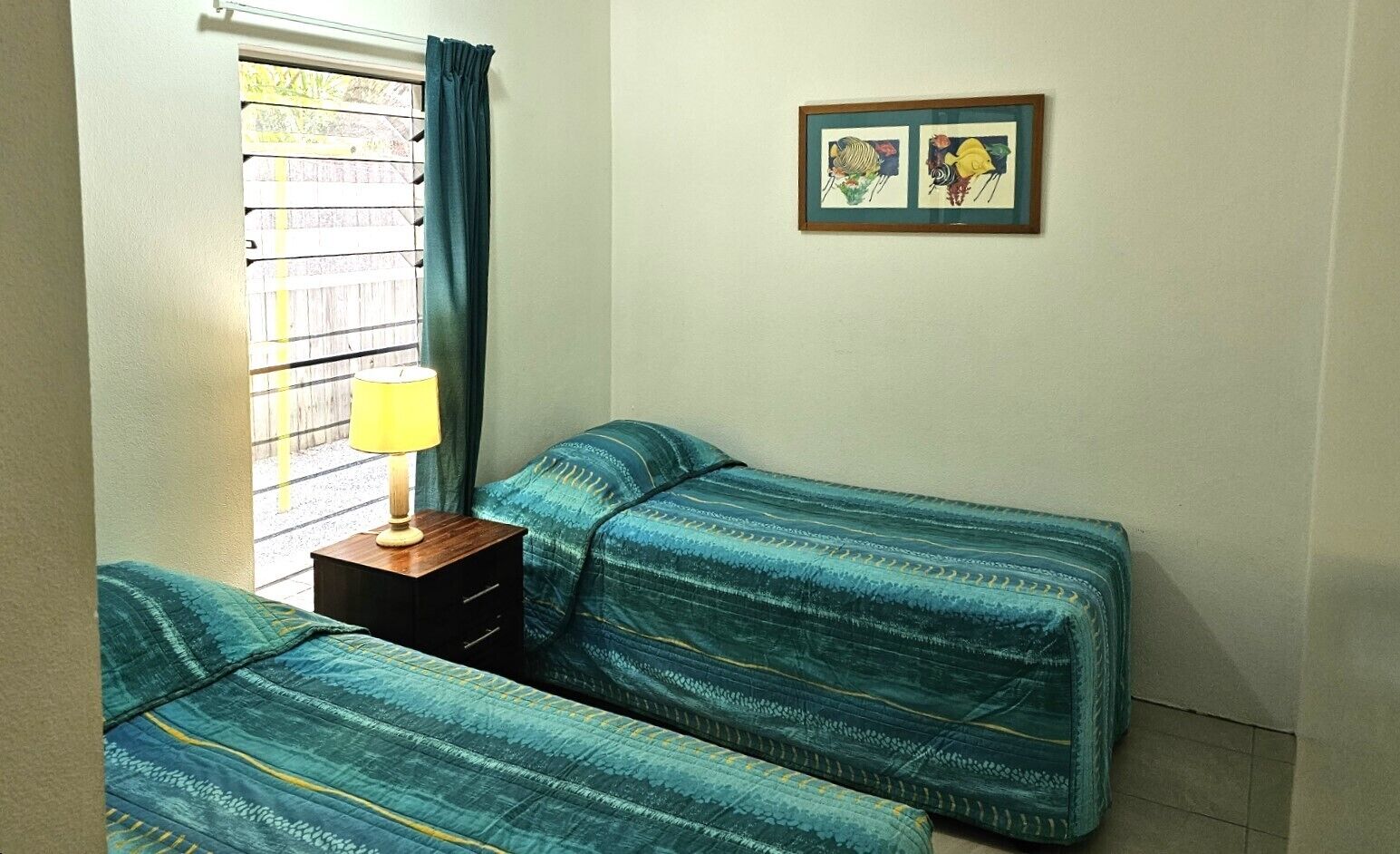 Una habitación pequeña y sencilla con dos camas individuales, una mesita de noche con lámpara y un cuadro en la pared.