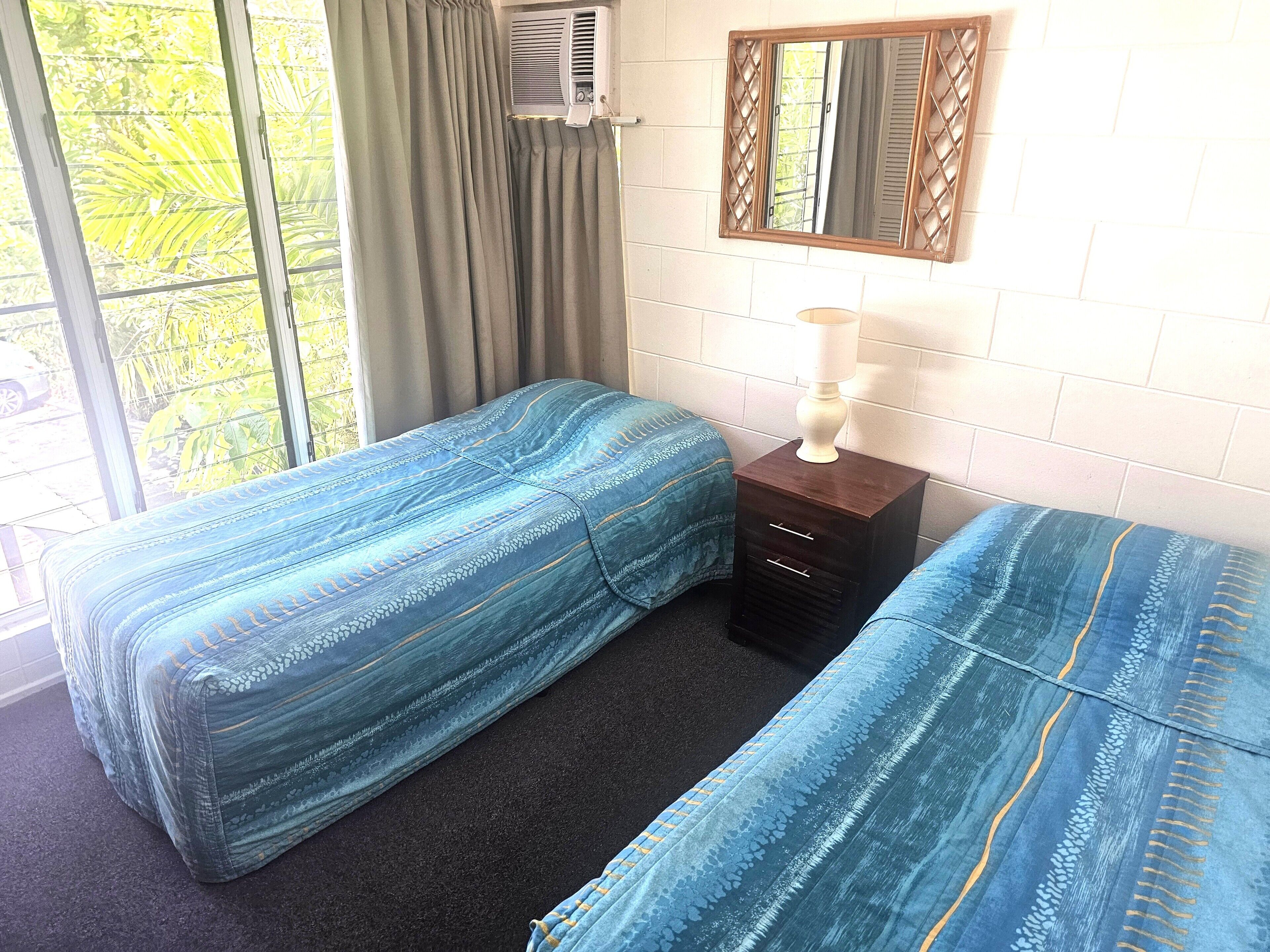 Une chambre avec deux lits, une petite table de chevet en bois avec une lampe, un miroir et un appareil de climatisation.