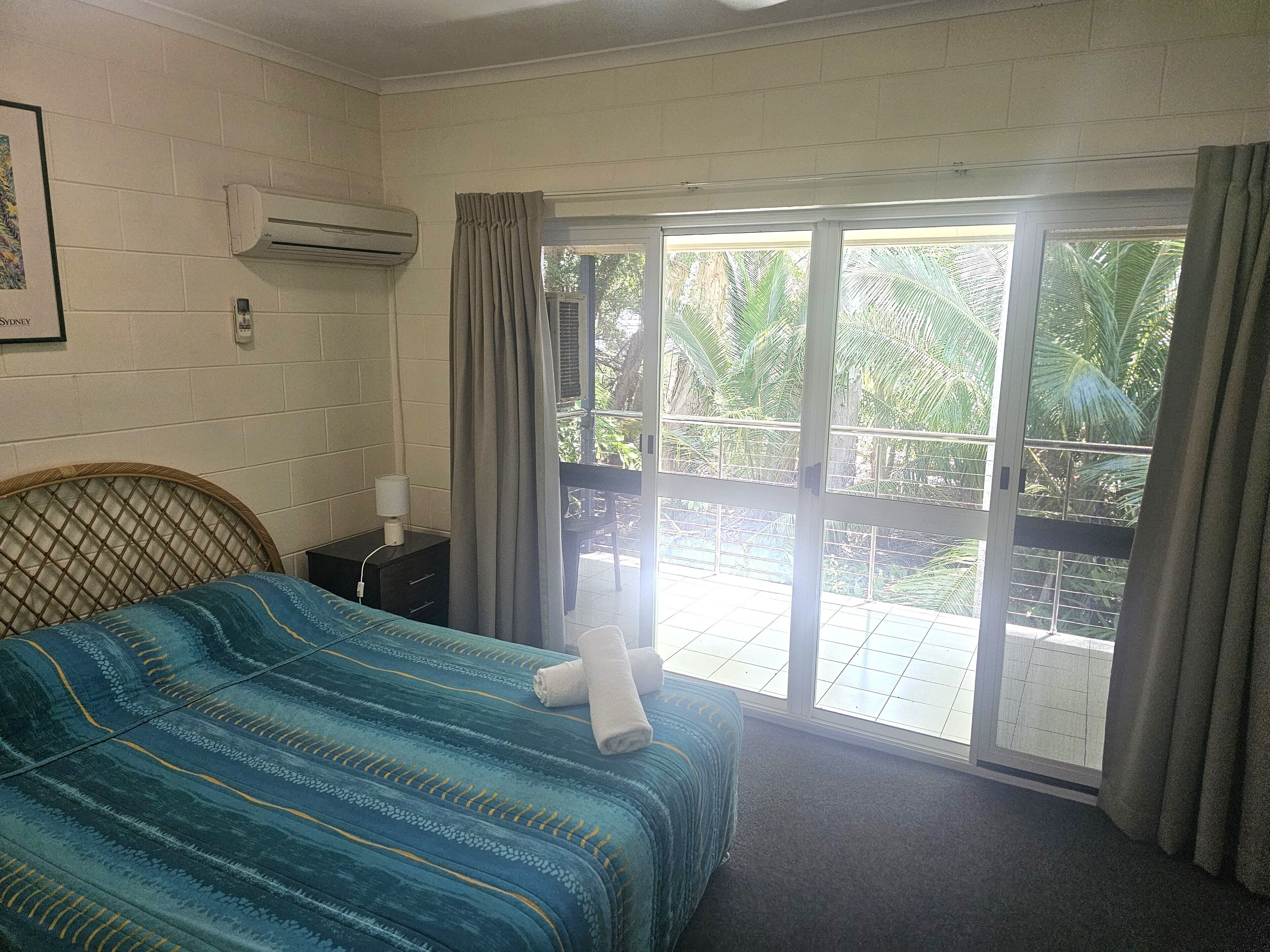 A hotel room with a bed, a nightstand, an air conditioner, and a view of the outdoors.