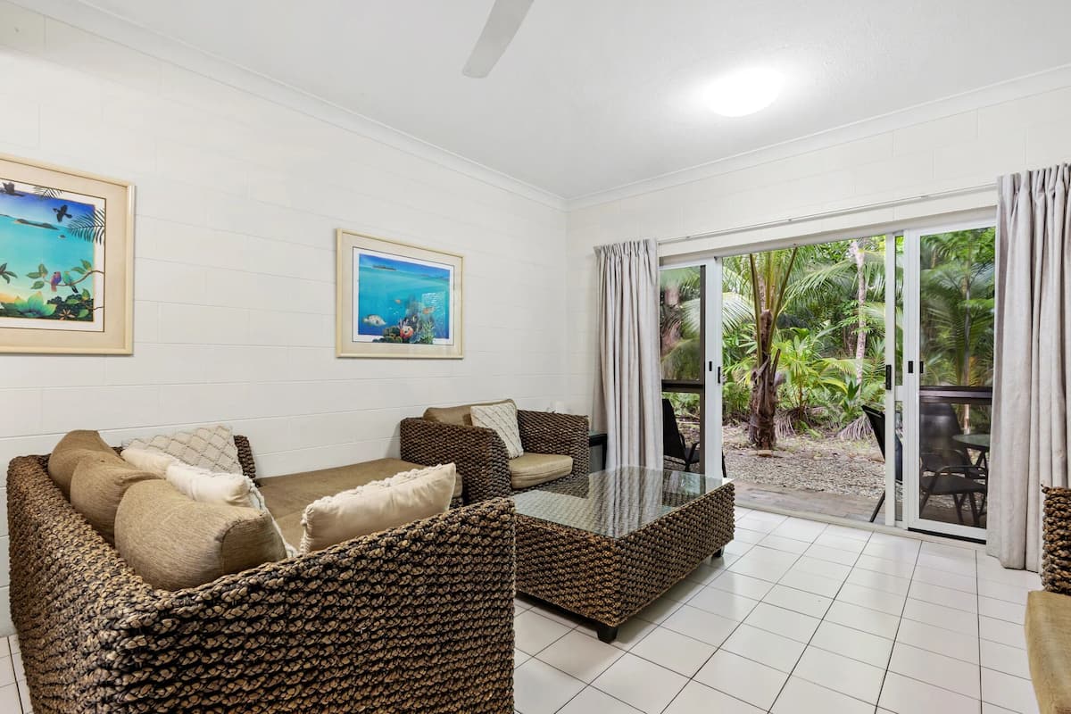 Habitación con sofá, una mesa de centro de vidrio y vista a un jardín tropical a través de una puerta corrediza de cristal.