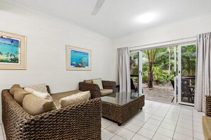 Une pièce comprenant un canapé, une table basse avec un plateau en verre et donnant sur un jardin tropical grâce à une porte-fenêtre coulissante.