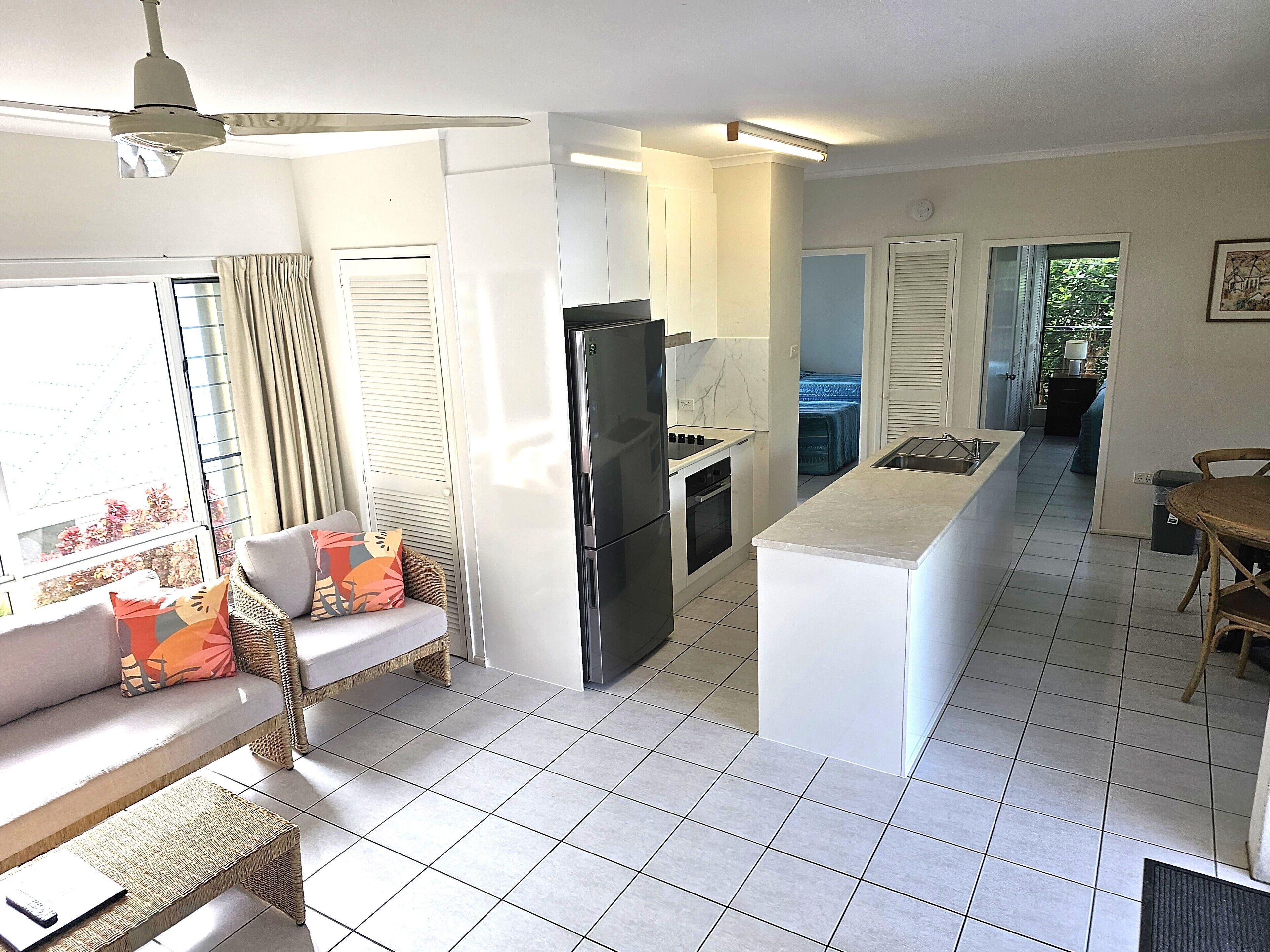 Cocina moderna con refrigerador, estufa y lavaplatos. Se observa un comedor con mesa y sillas. Un sofá con cojines está cerca de una ventana.